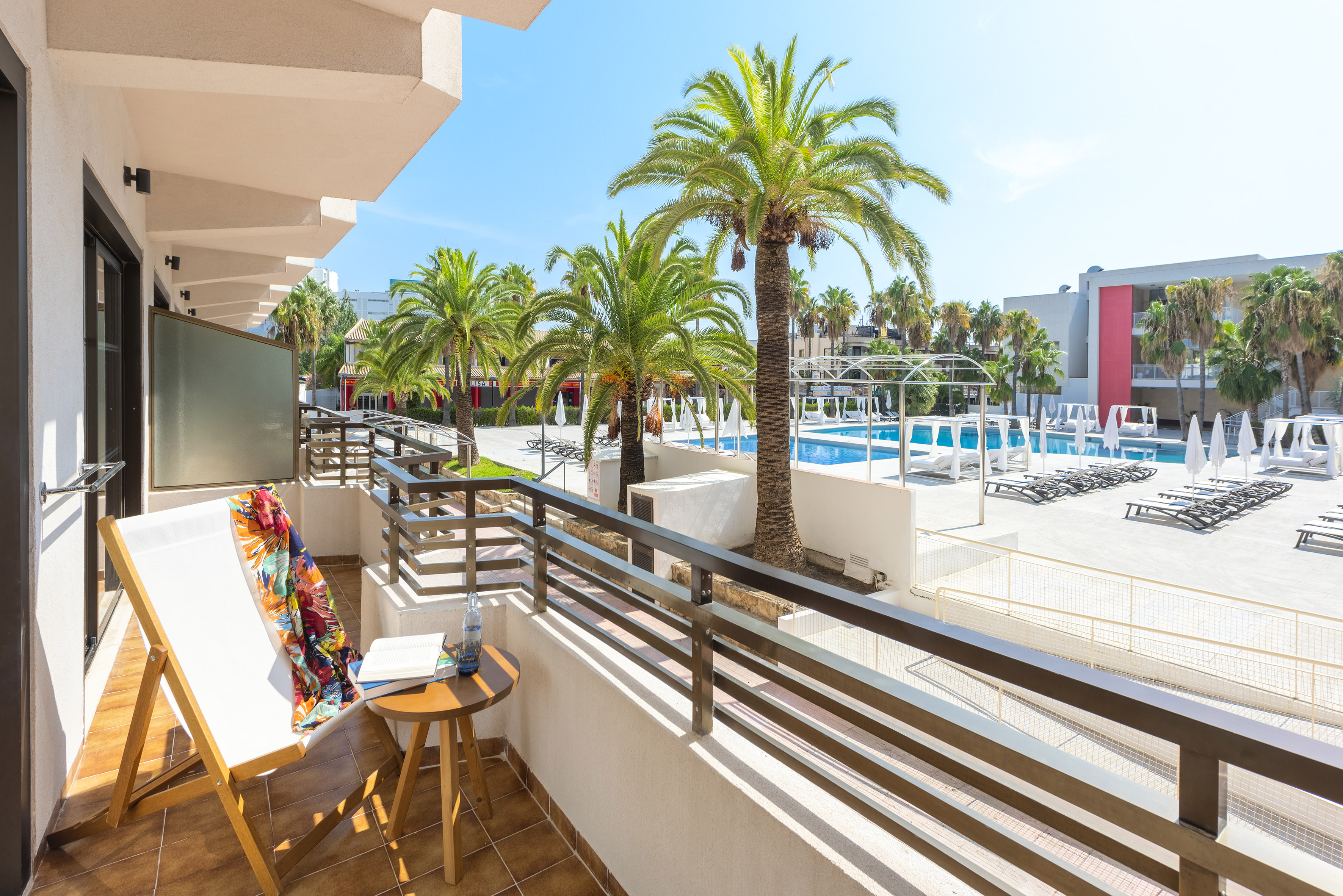 Hotel balcony overlooking a sunny pool area with palm trees and sun loungers.