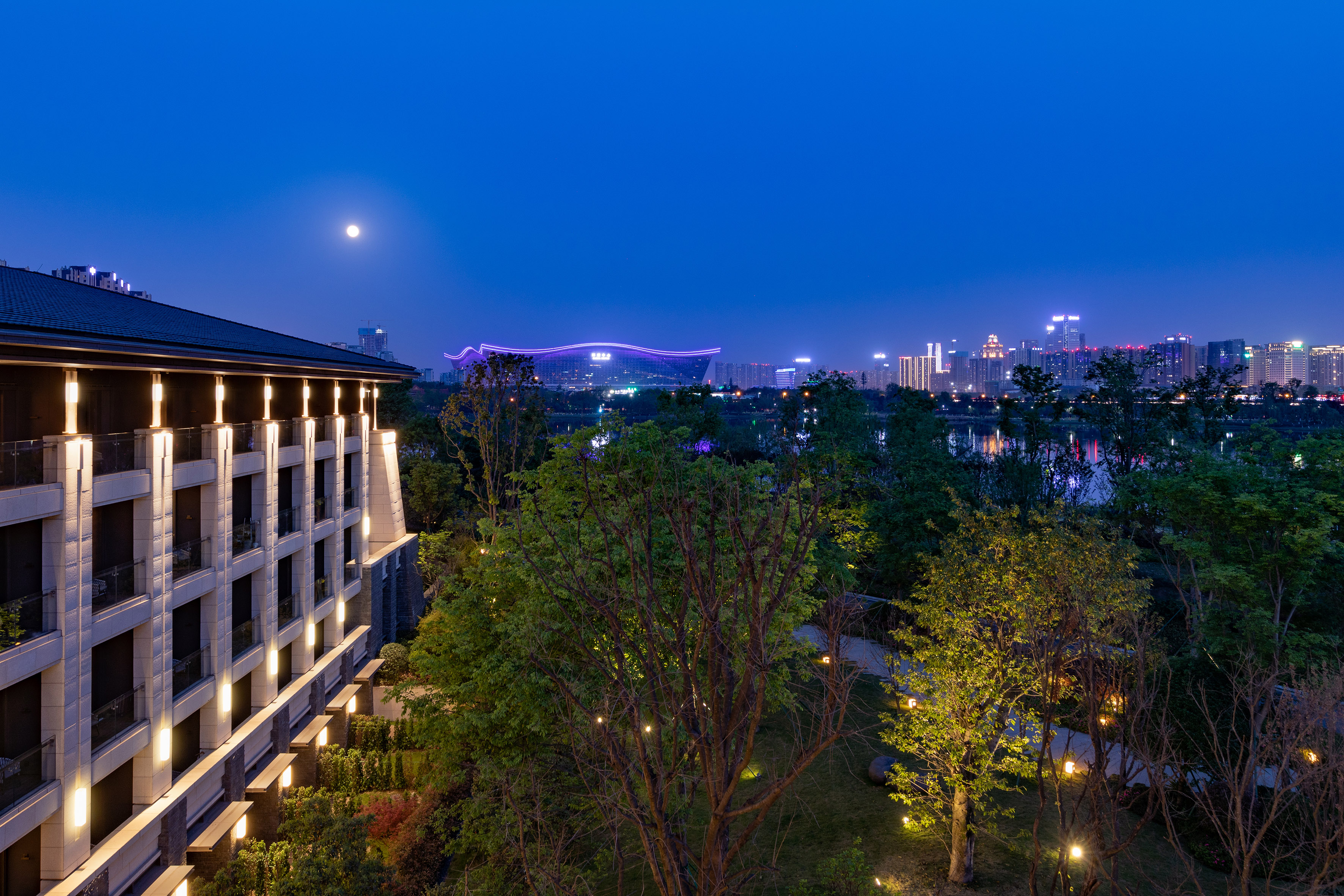 Illuminated modern building, full moon, city skyline, and waterfront at night.