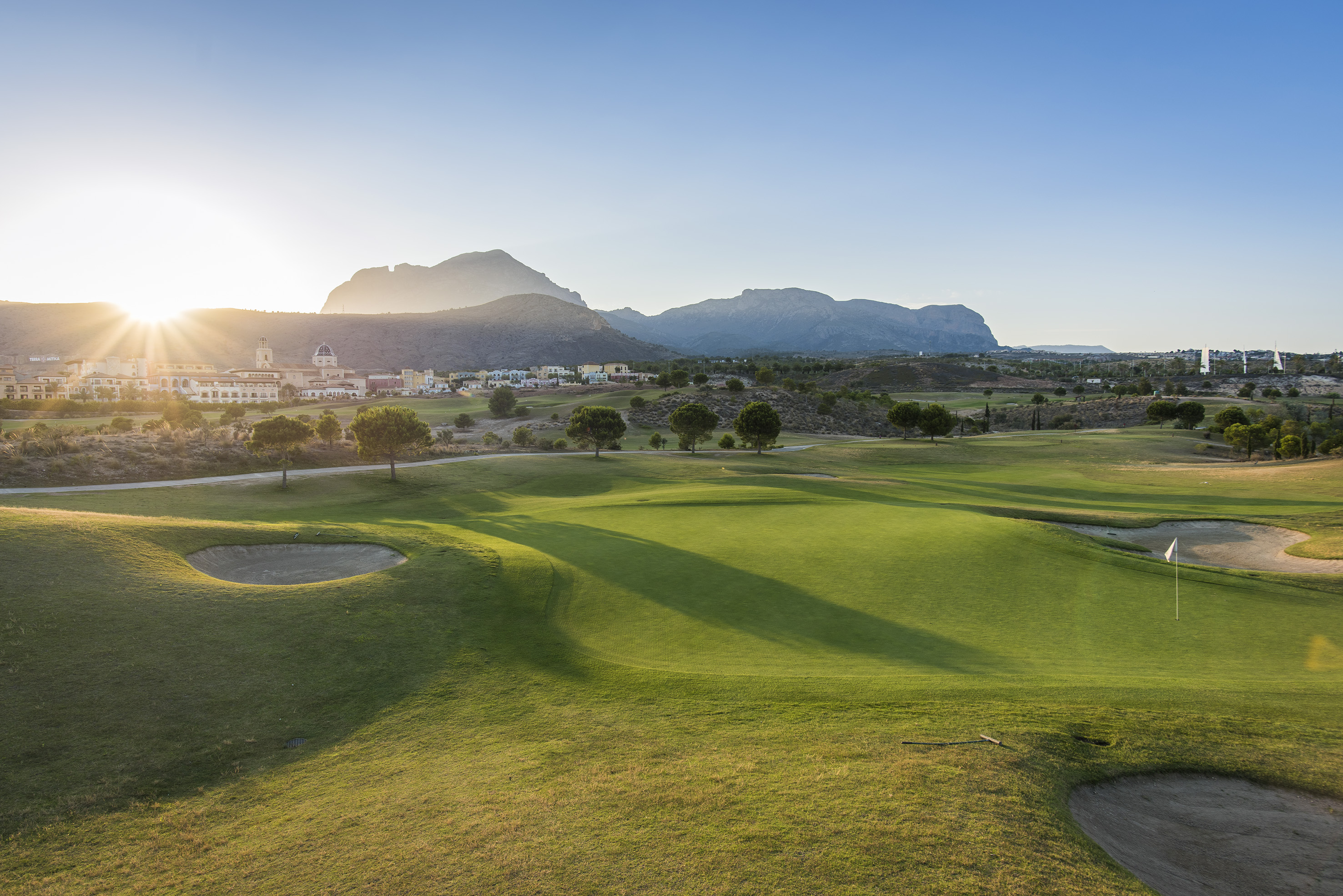 Golf course green, bunkers, distant village, and mountains under a sunset sky.