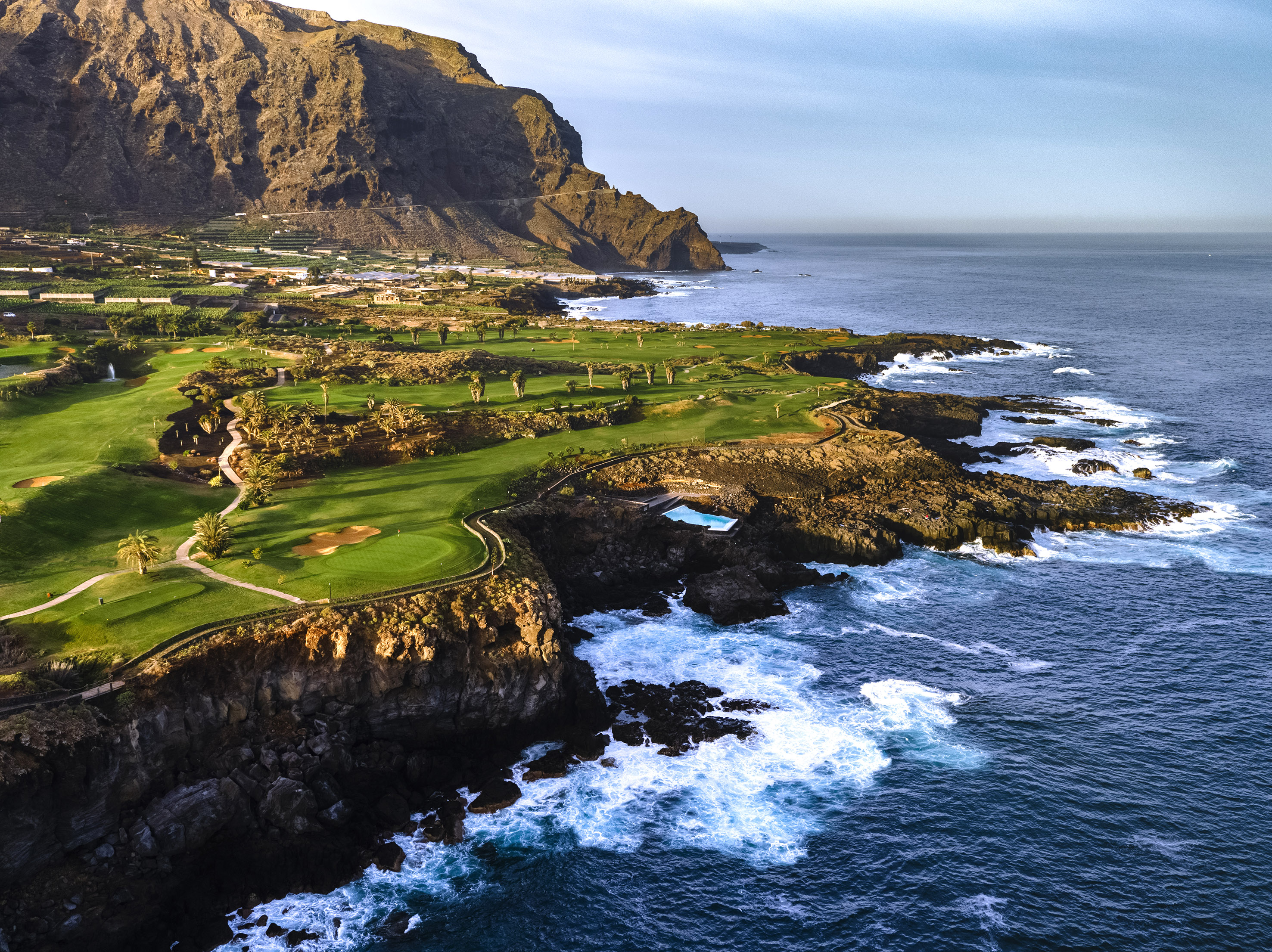 Oceanfront golf course on rocky cliffs, crashing waves, and dramatic mountains.