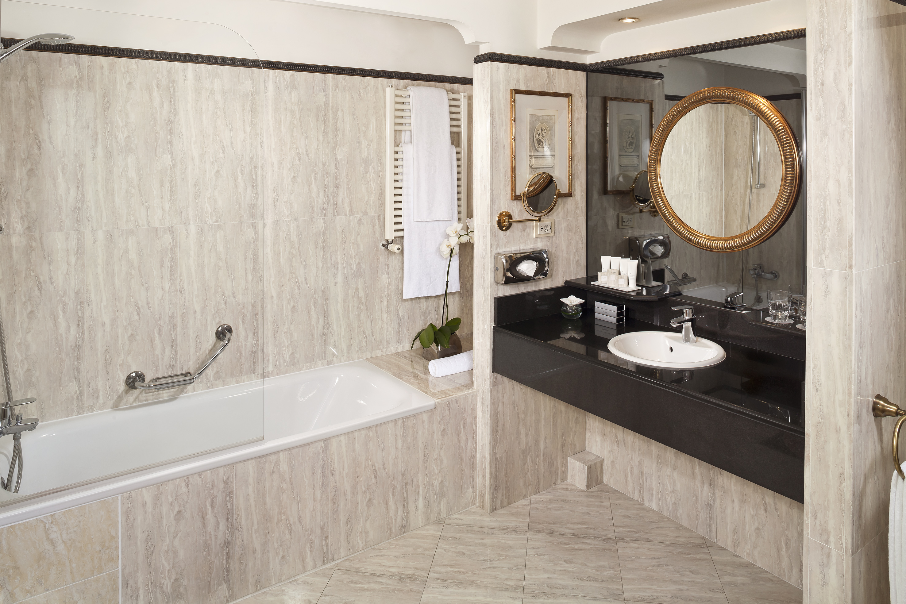 Hotel bathroom featuring marble walls, bathtub, black vanity, and round gold mirror.