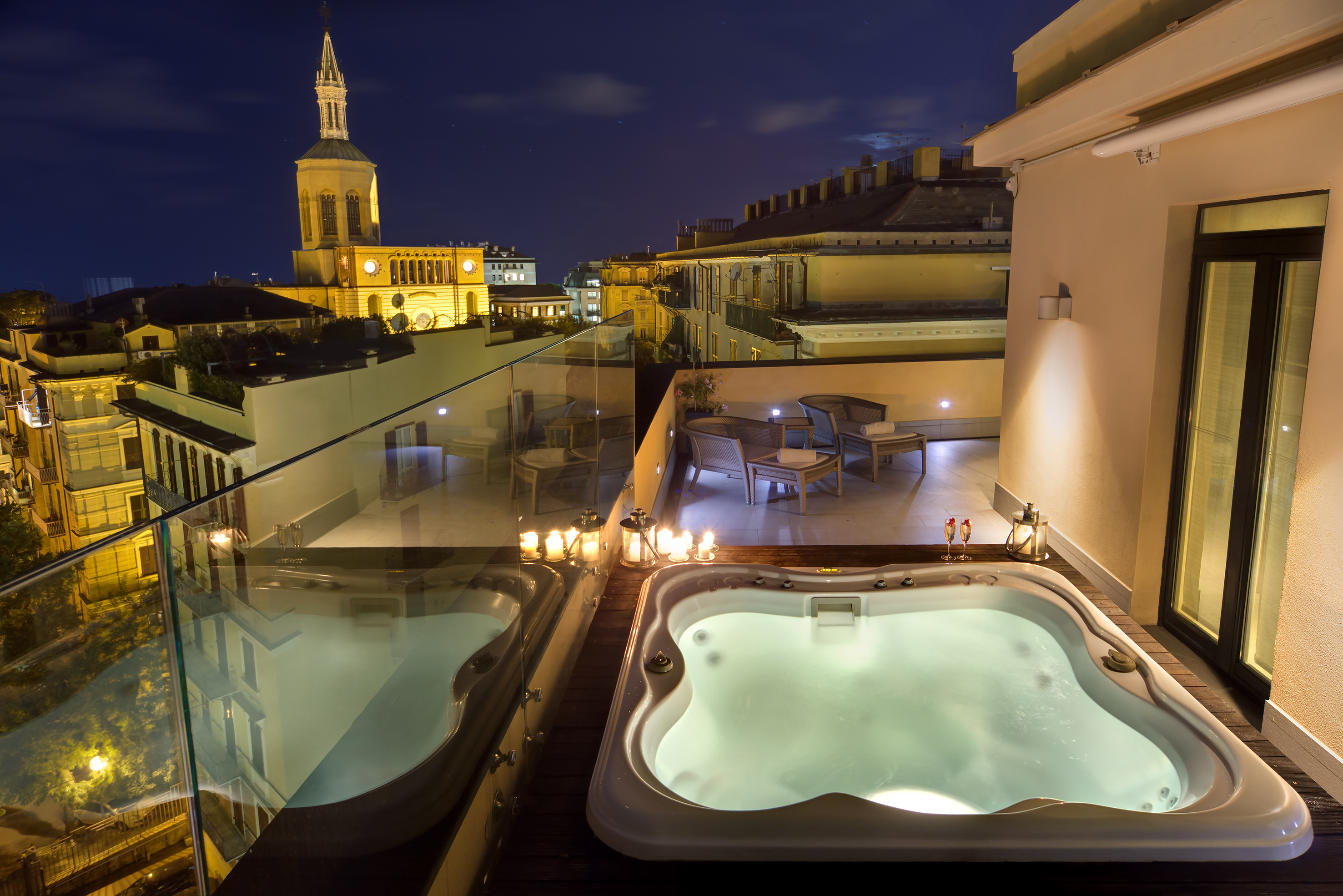 Nighttime rooftop terrace with two jacuzzis, cityscape, and illuminated church tower.