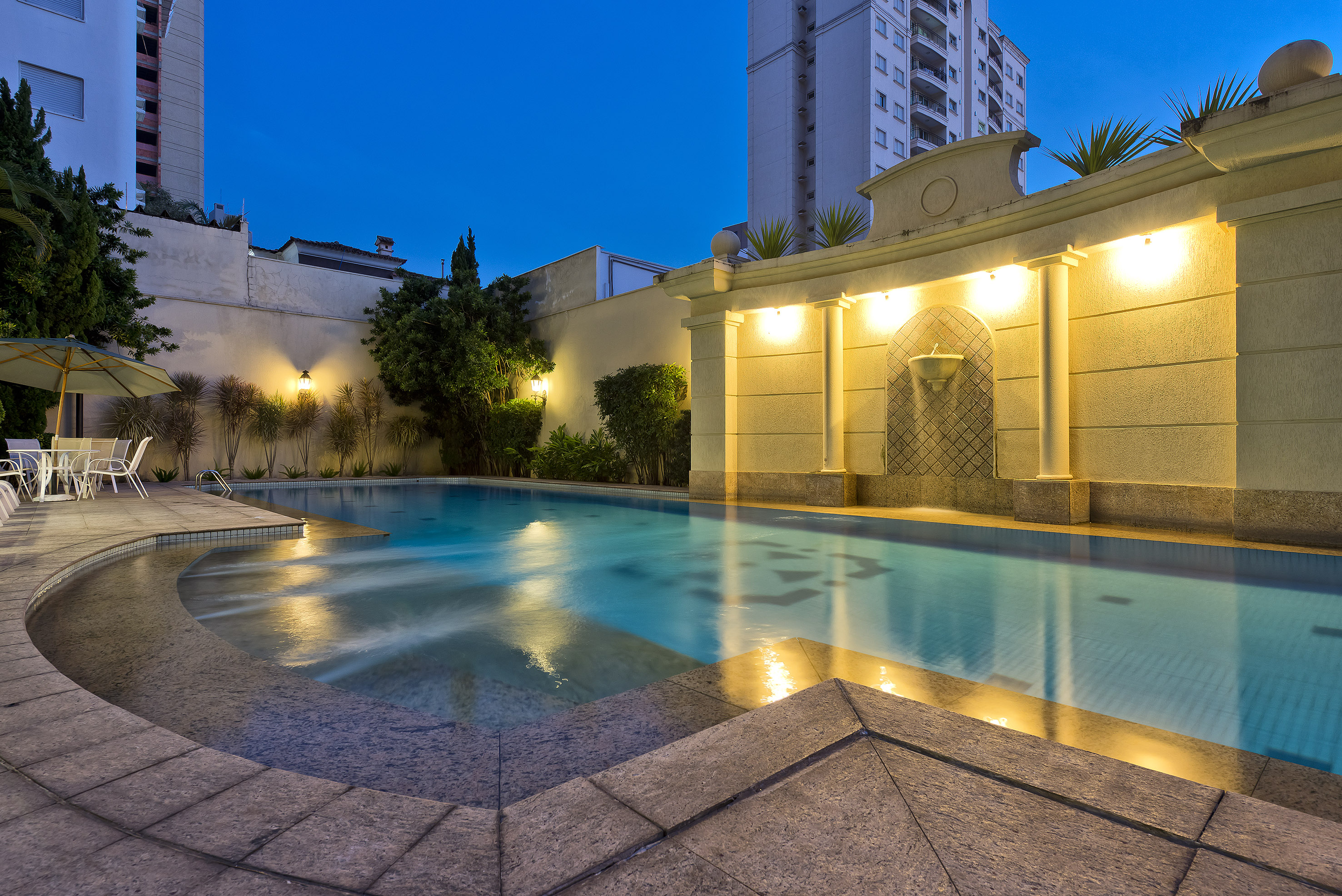 Illuminated outdoor pool at night with fountain, patio, and building.