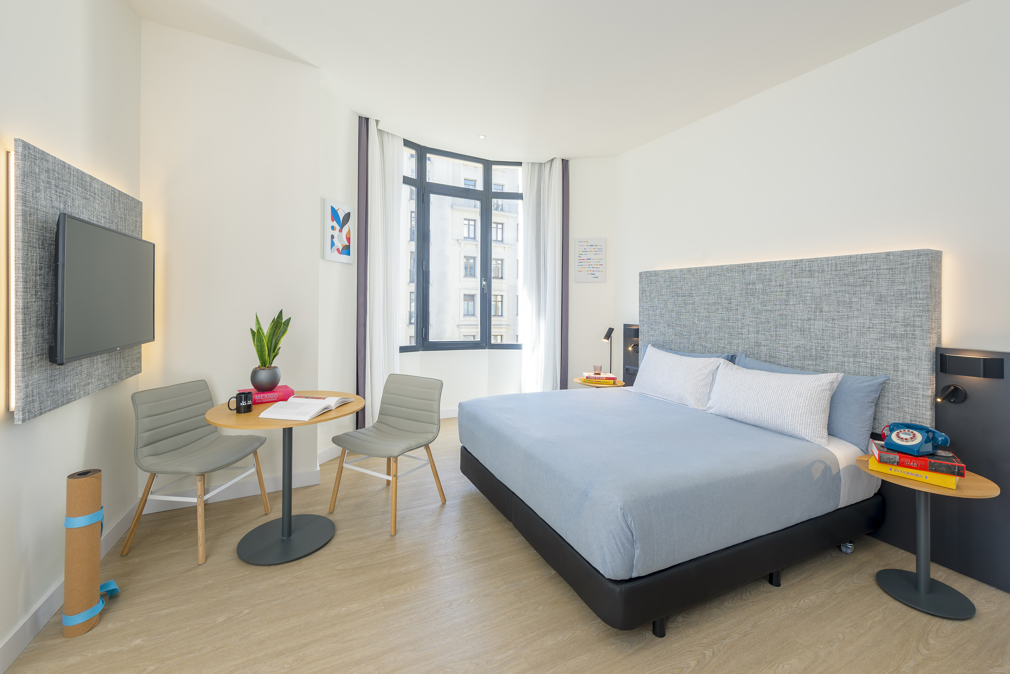 Bright modern hotel room with queen bed, bay window, TV, and work table.