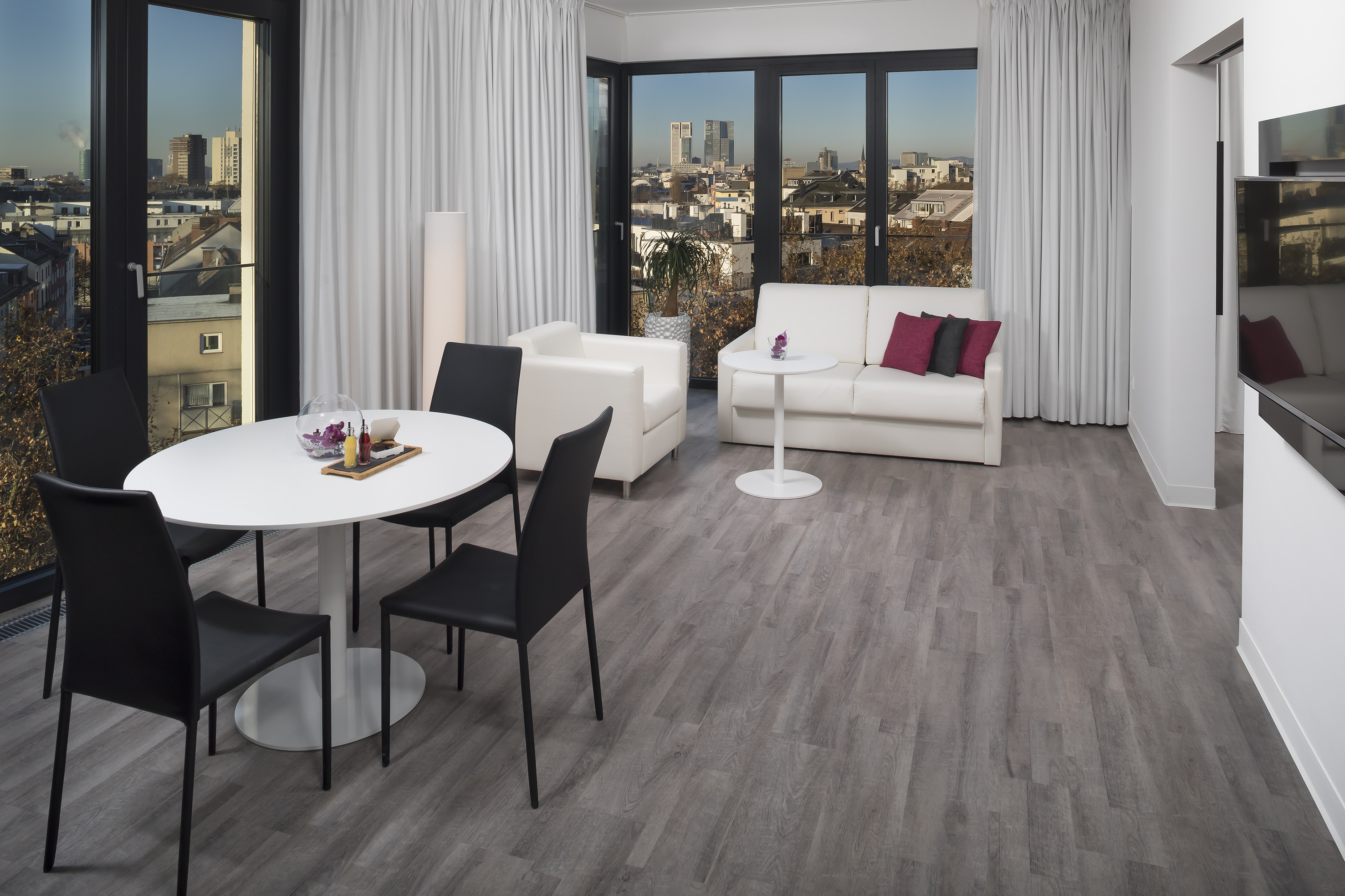 Modern hotel suite, city view, dining area, white sofa, grey floor.