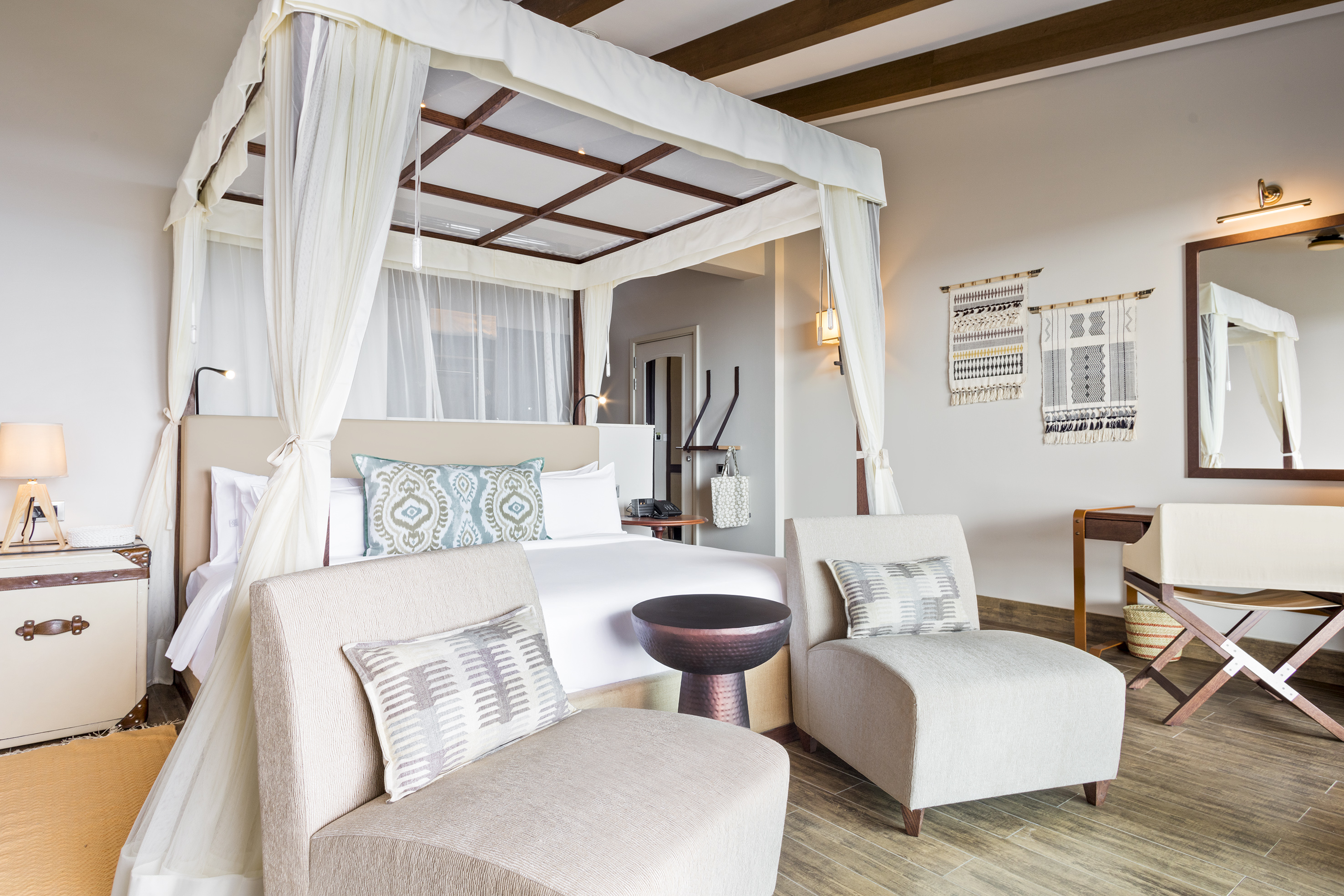 Stylish hotel room with white four-poster bed, two armchairs, and woven art.
