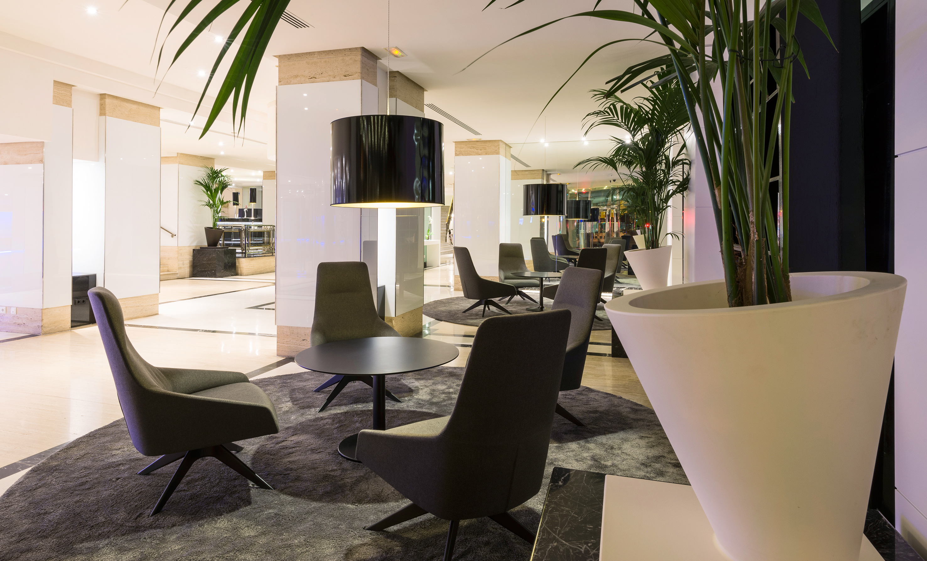 Modern hotel lounge with gray chairs, round tables, pendant lights, and potted plants.