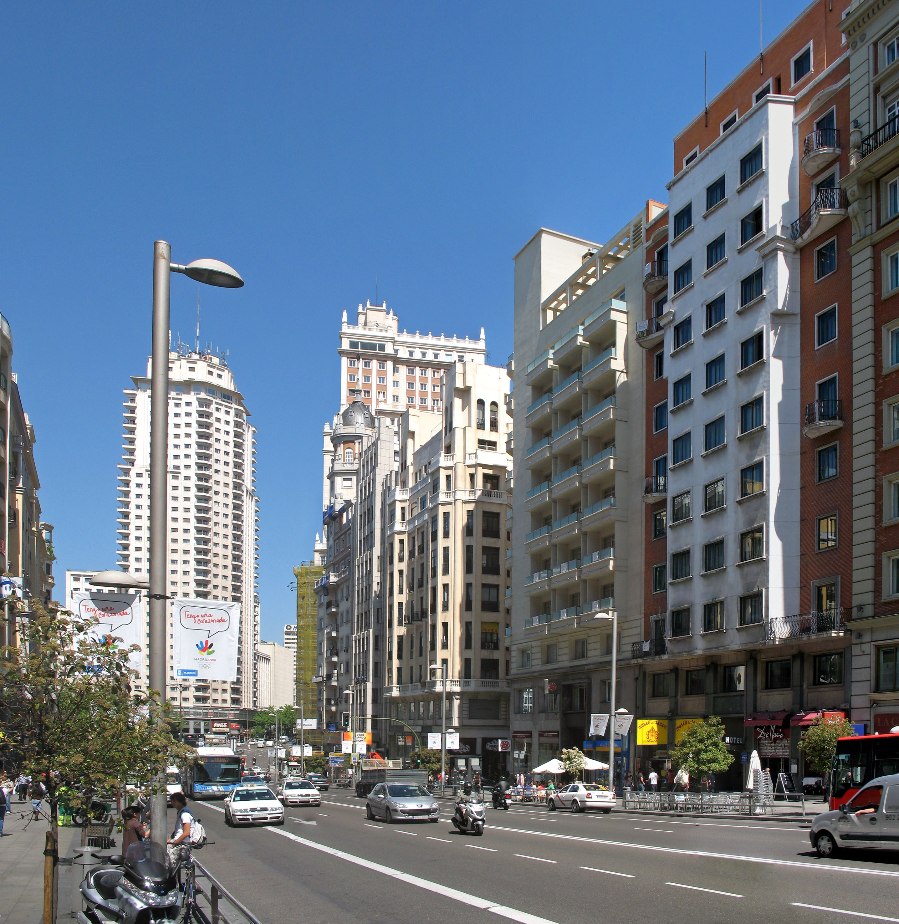 Madrid city street with buildings, cars, pedestrians, and a clear blue sky.