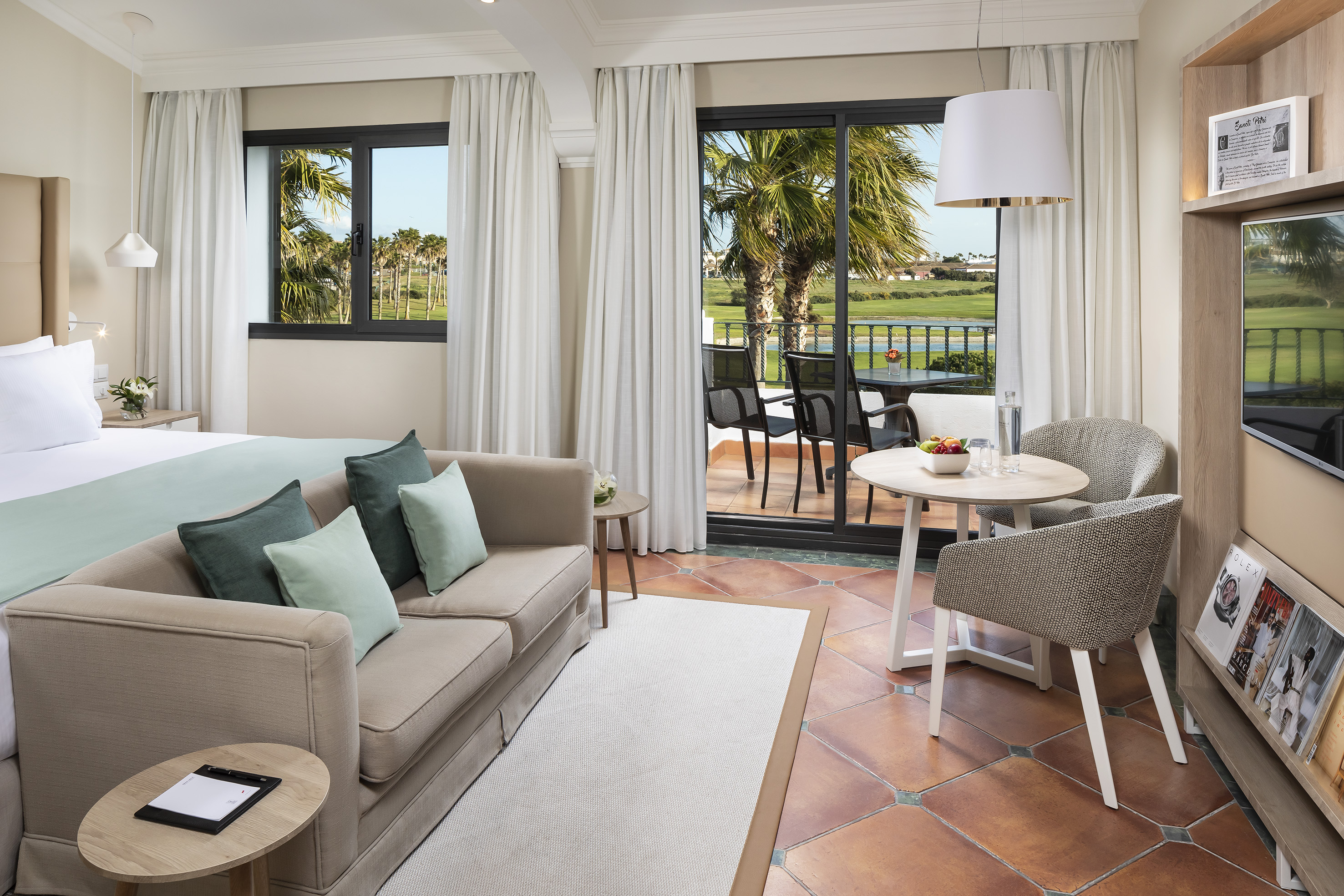 Modern hotel suite with golf course view, balcony, sofa, and dining area.