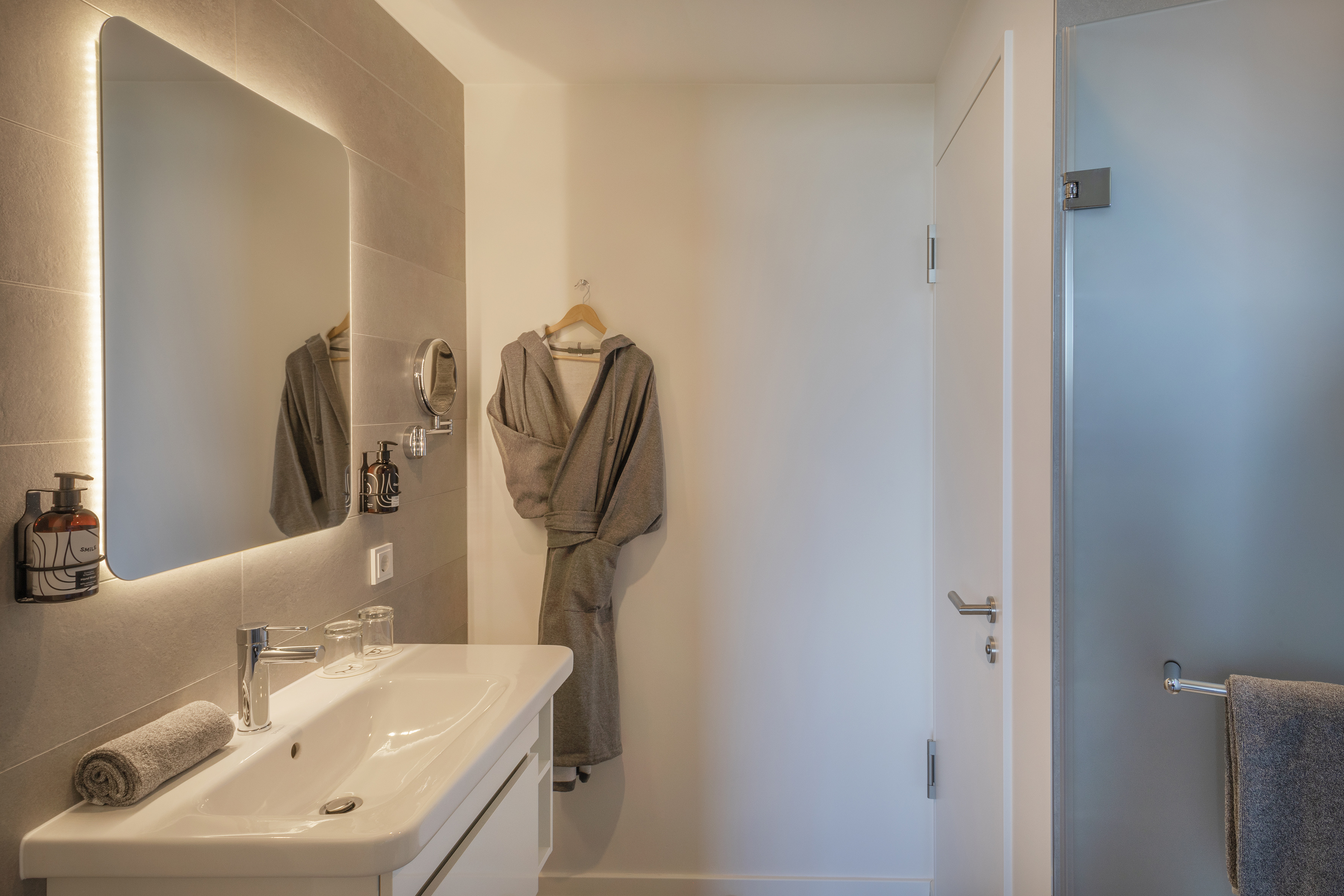 Contemporary bathroom with illuminated mirror, white vanity, and hanging bathrobe.