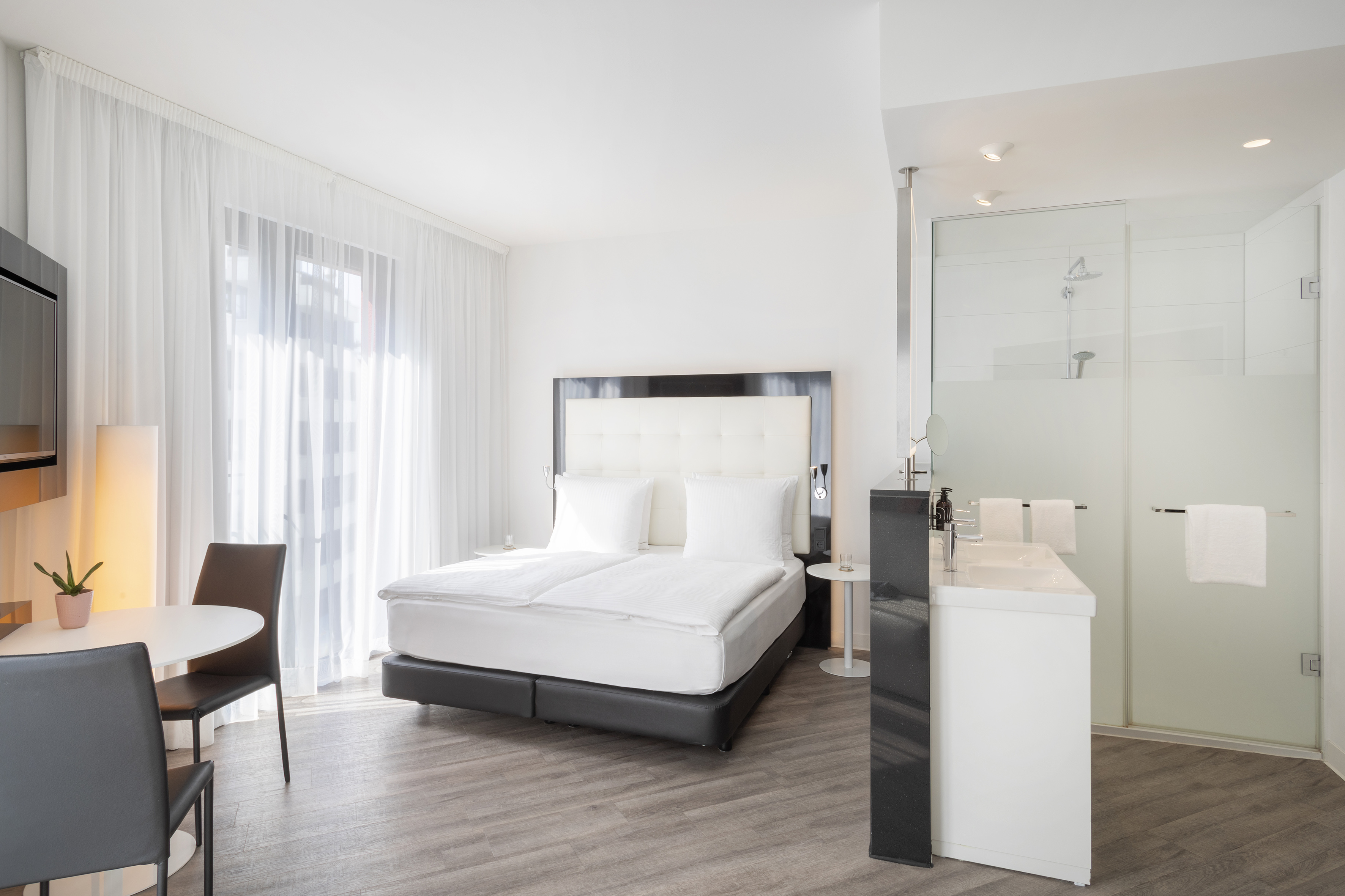 Bright modern hotel room with large bed, dining space, and glass shower.