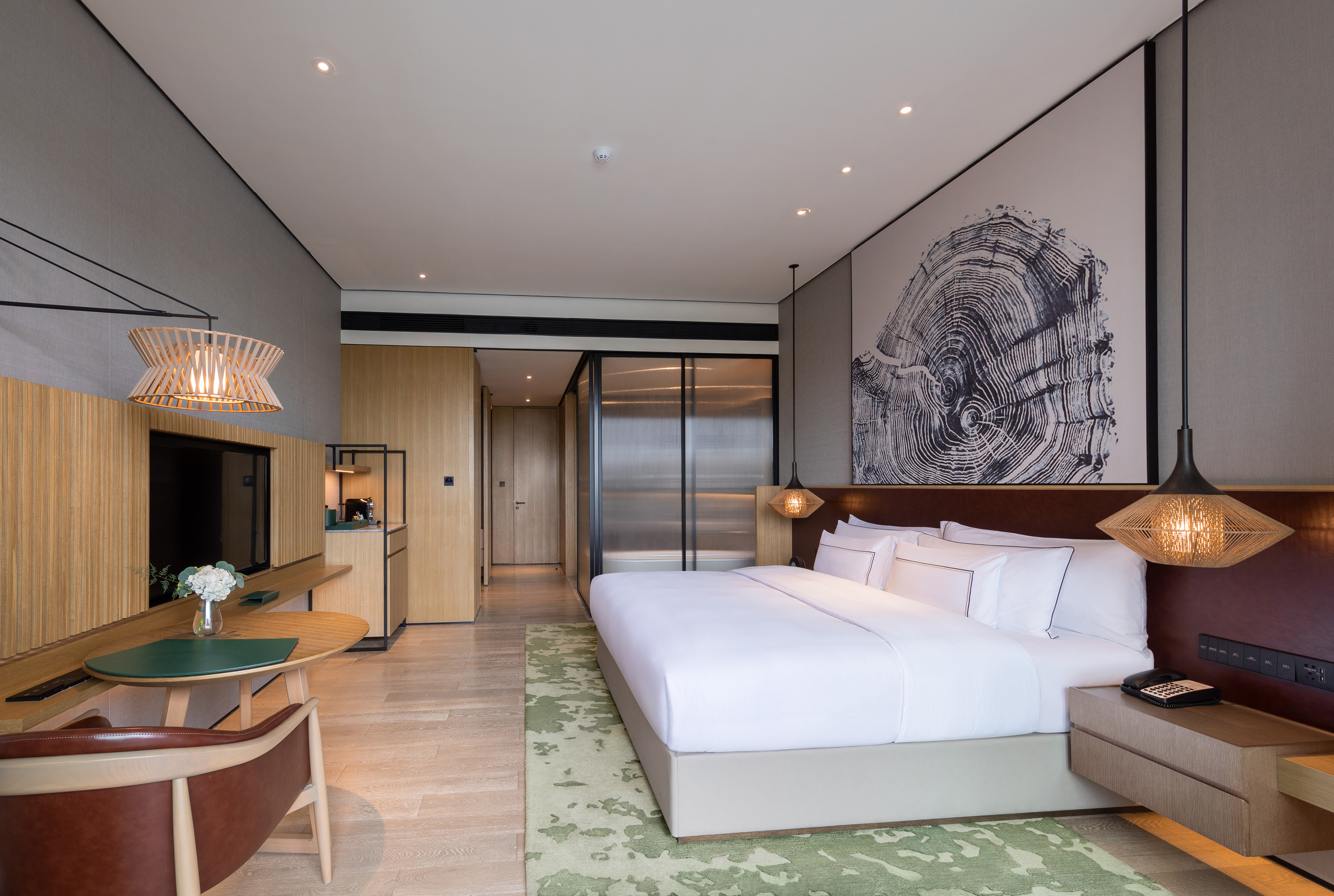 Modern hotel room with king bed, wood decor, green rug, and tree ring artwork.