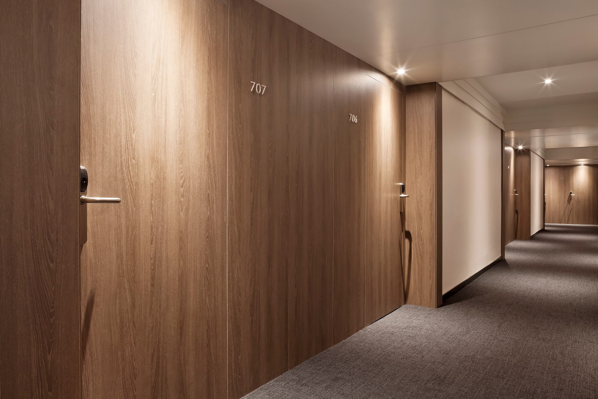 Modern hotel hallway with wooden room doors, gray carpet, and bright lighting.