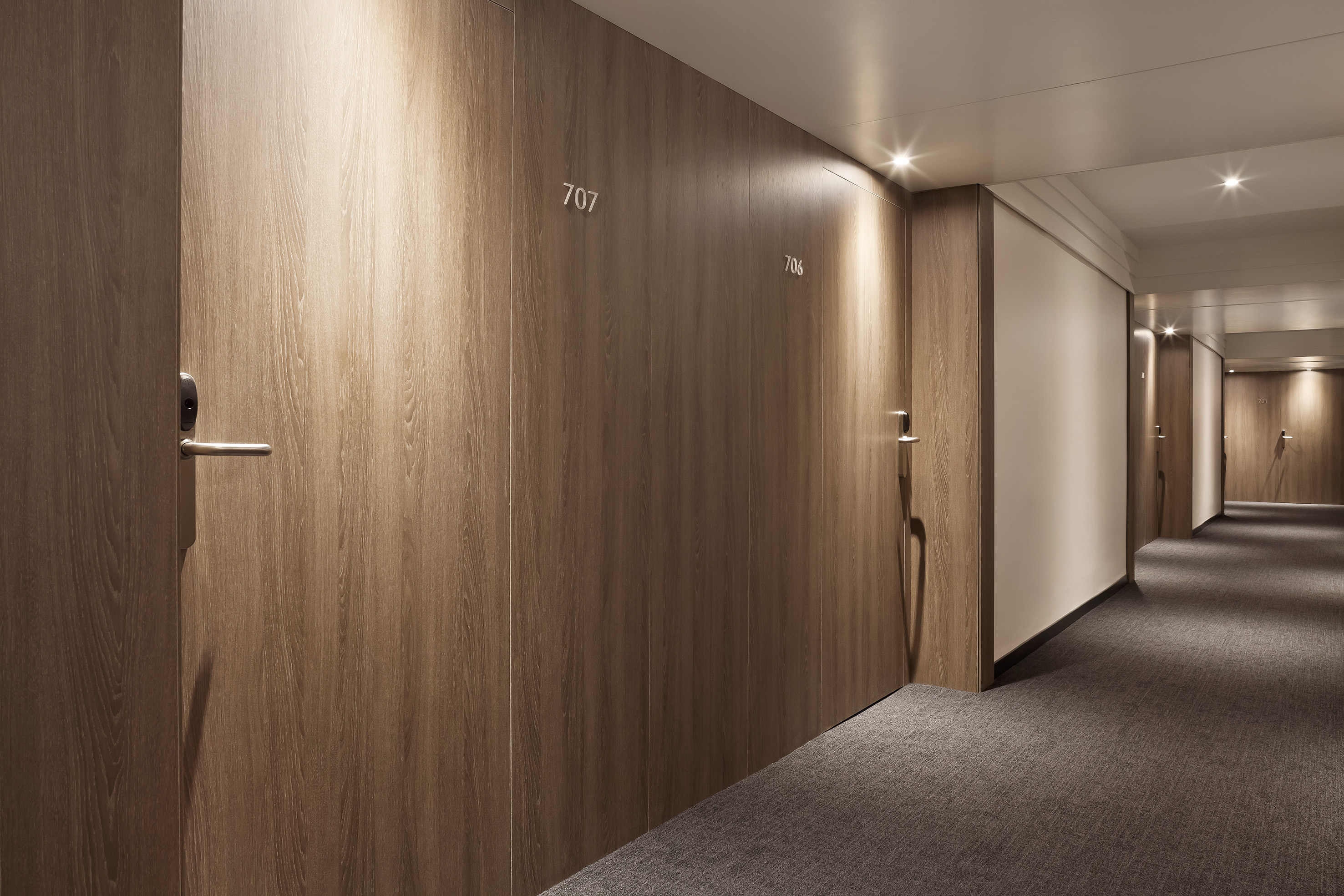 Modern hotel hallway with wooden room doors, gray carpet, and bright lighting.