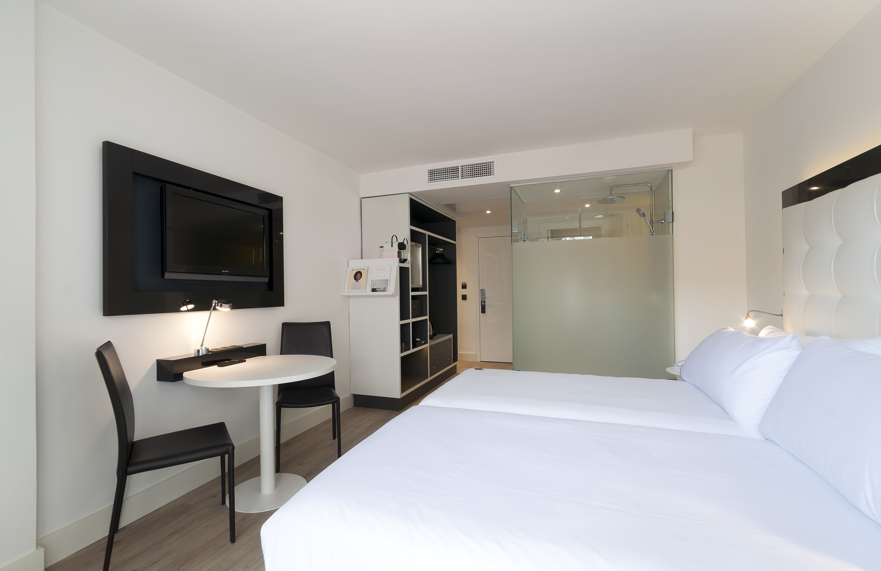 Modern hotel room with twin beds, wall TV, small table, and glass shower.