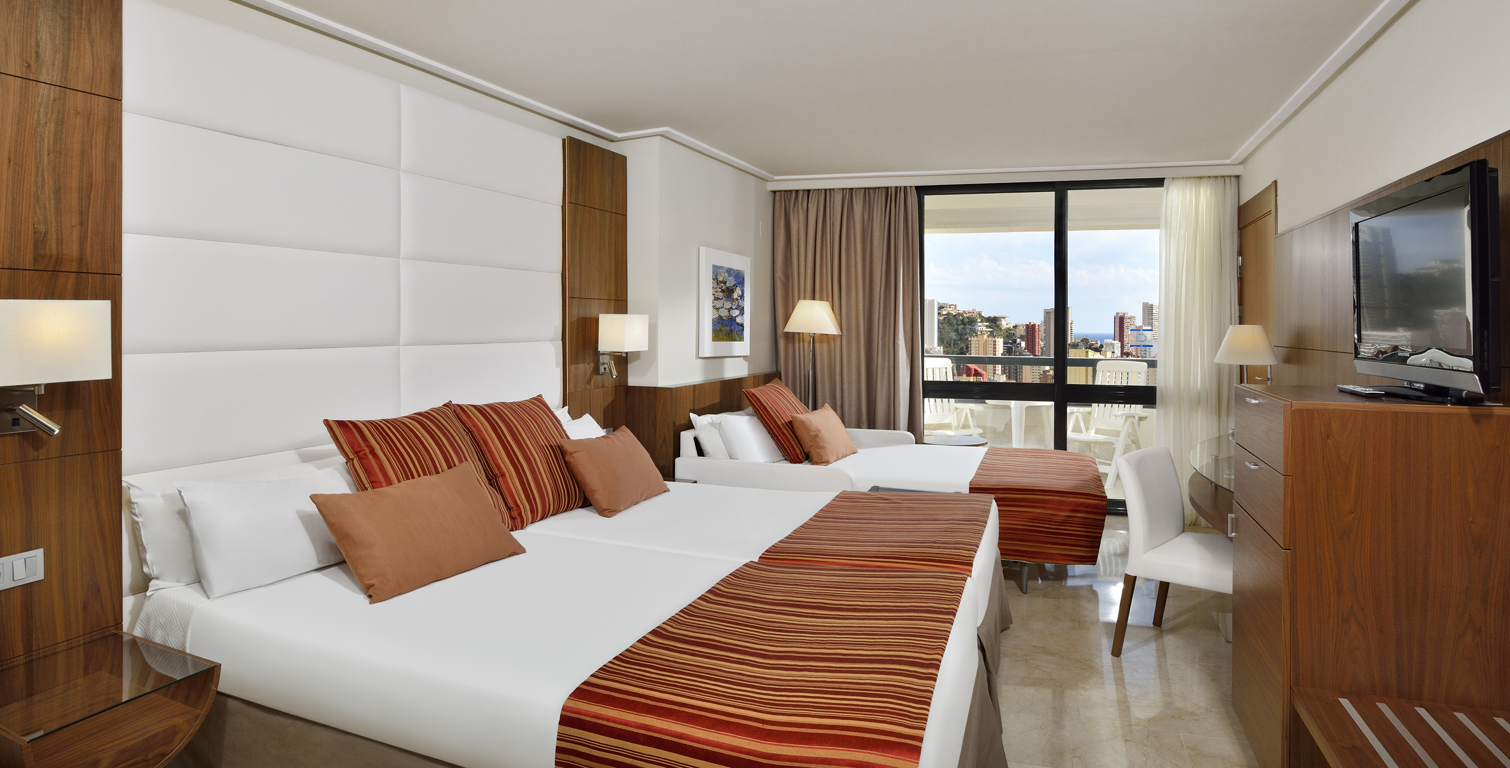 Modern hotel room with twin beds, sofa bed, balcony, and city-sea view.