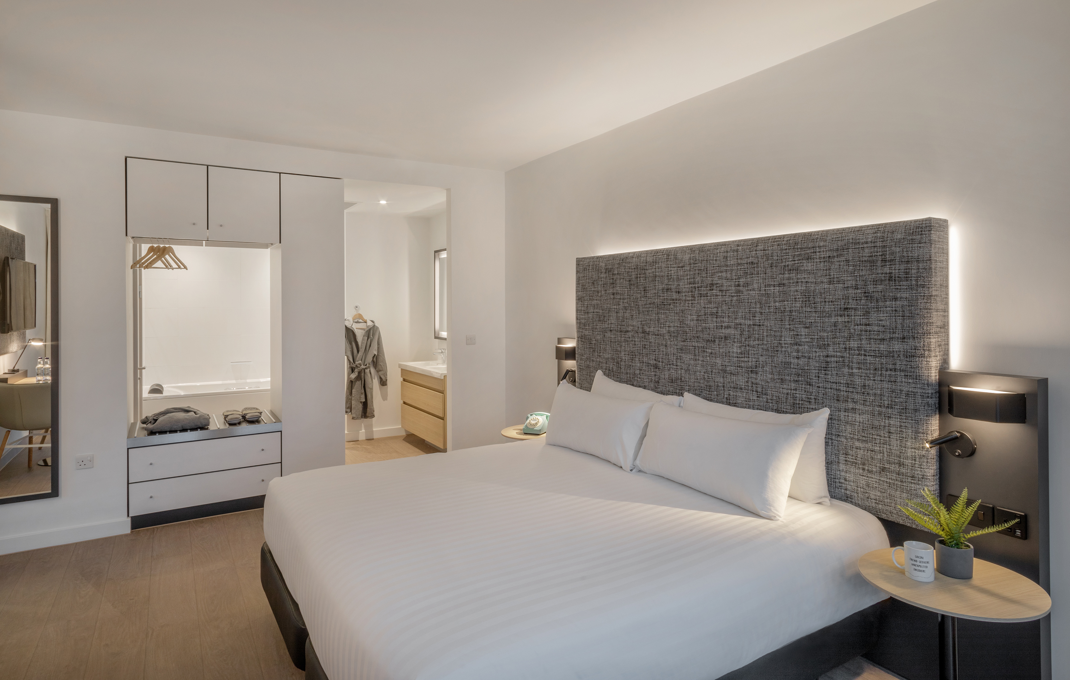 Modern hotel bedroom with white bed, grey headboard, and open bathroom.