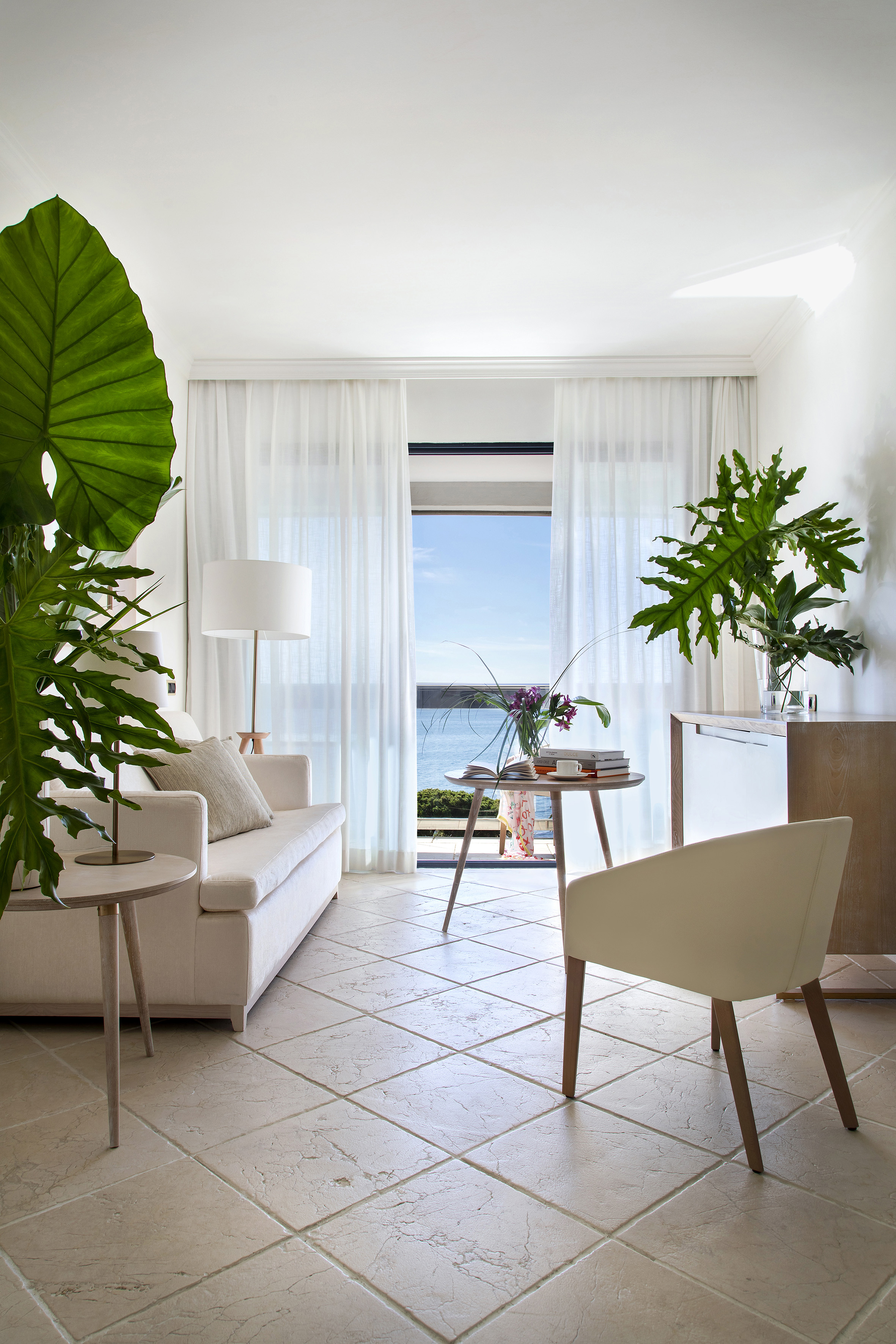 Modern living room with sea view, beige sofa, lush plants, neutral decor.