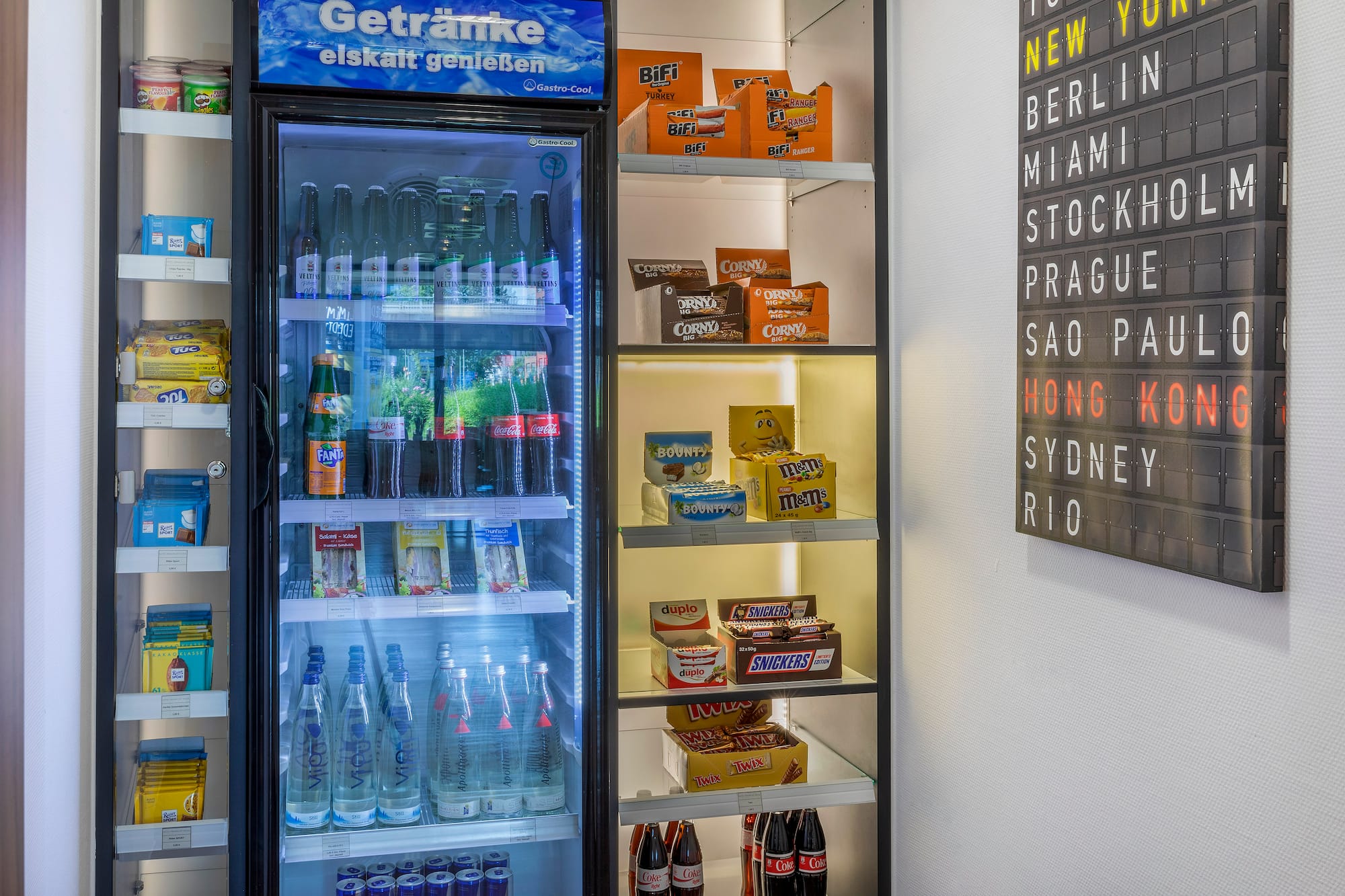 Hotel mini-bar fridge with cold drinks, snacks, and a city destination board.