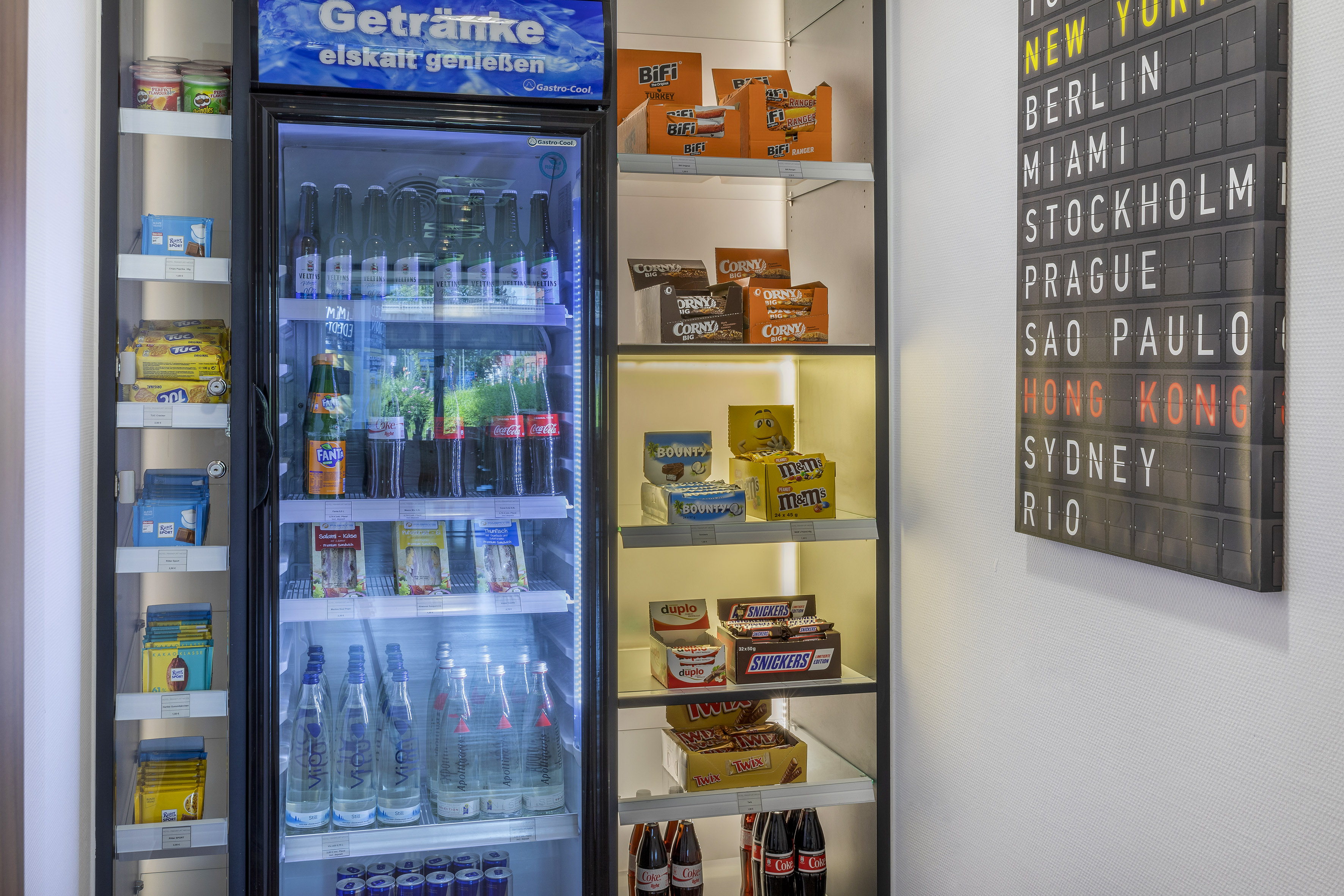 Hotel mini-bar fridge with cold drinks, snacks, and a city destination board.