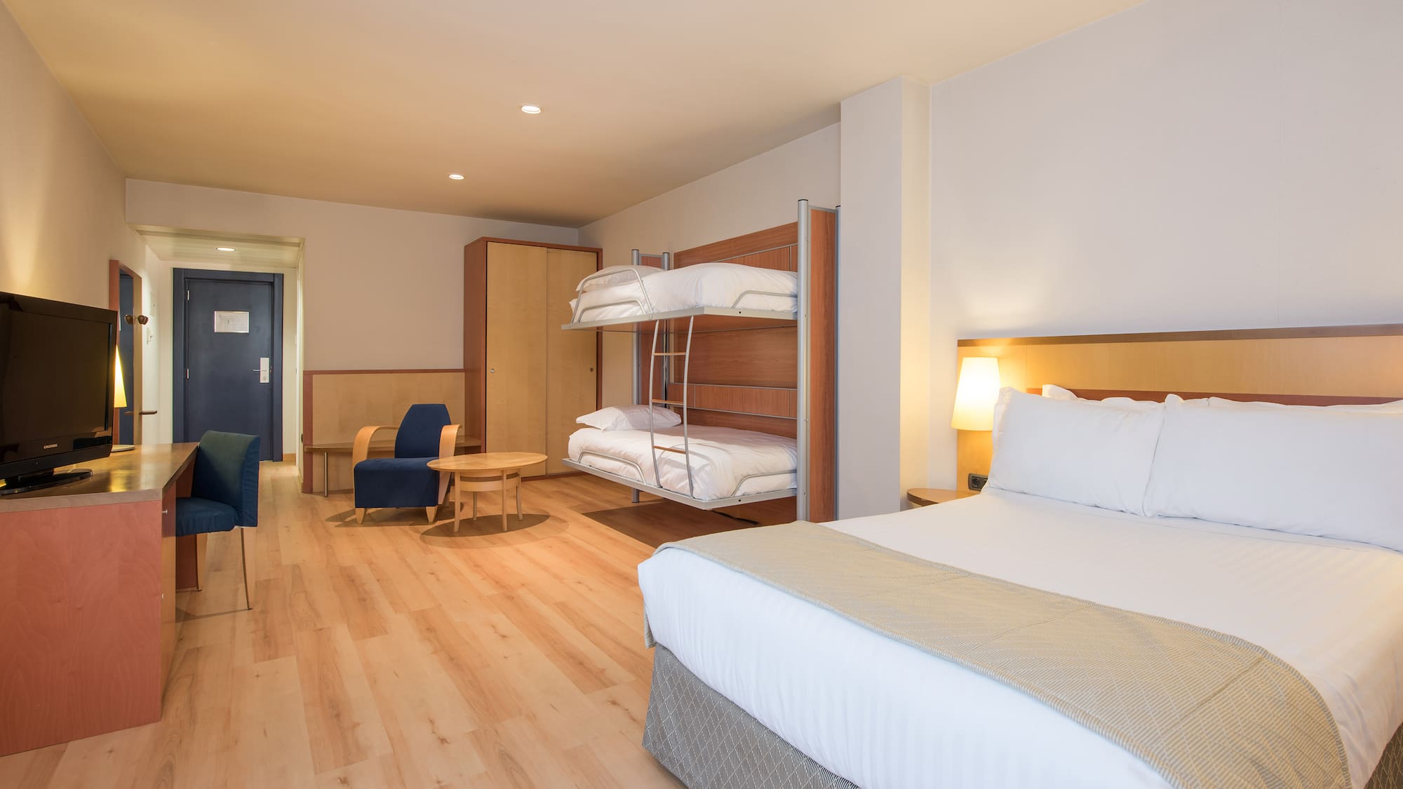 Modern hotel family room with king bed, bunk beds, desk, and armchair.