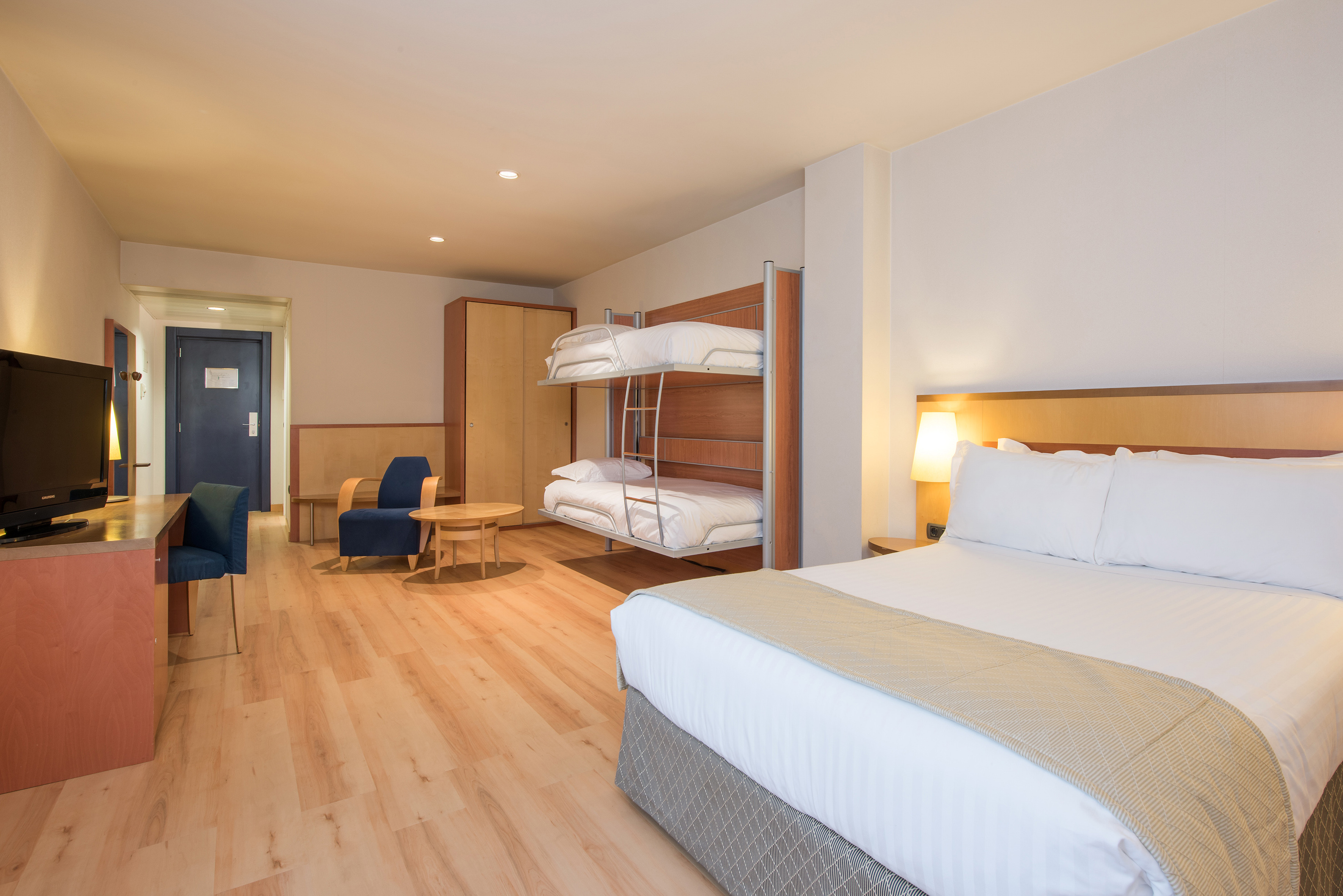 Modern hotel family room with king bed, bunk beds, desk, and armchair.