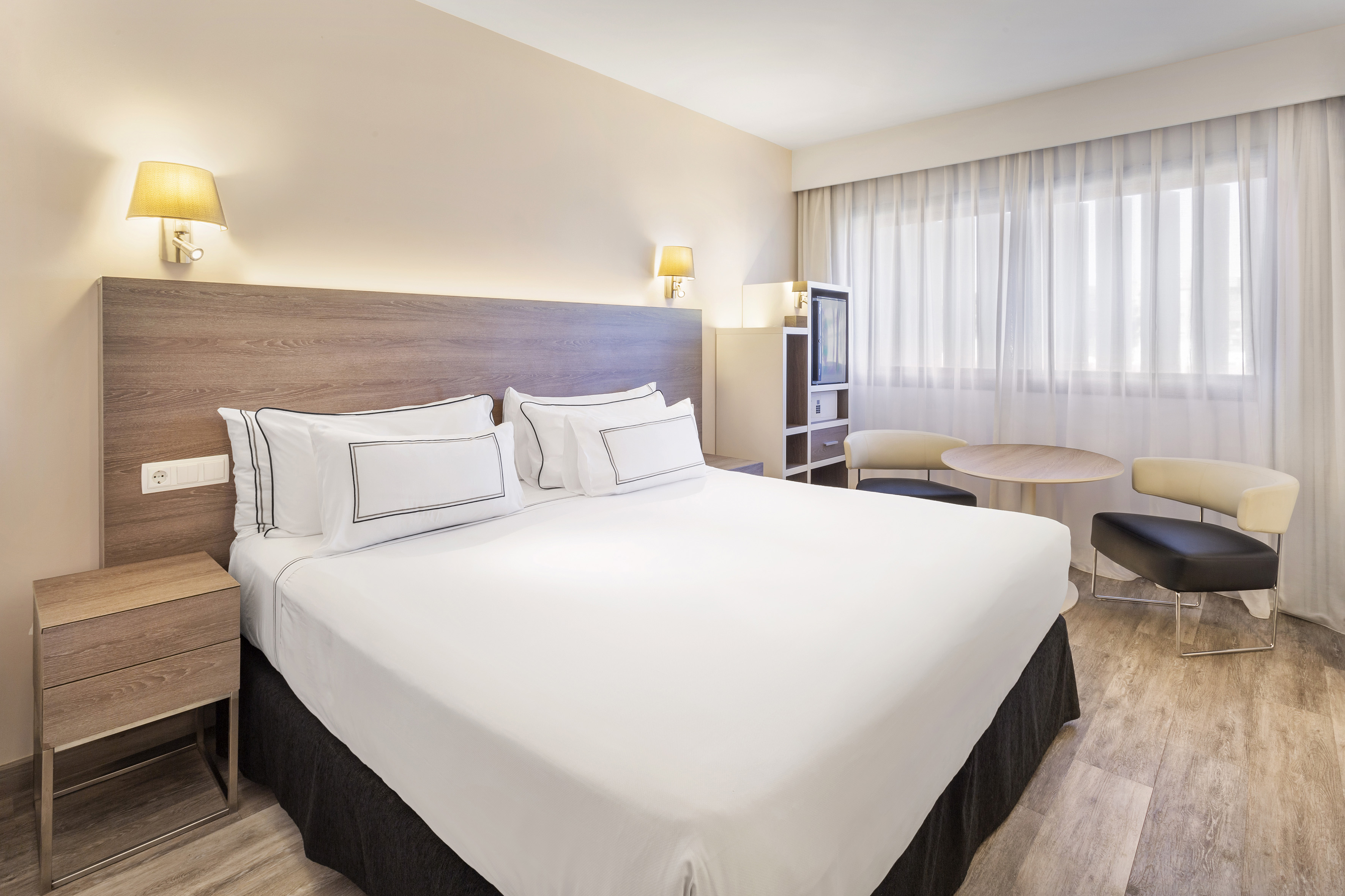 Modern hotel room with queen bed, wooden headboard, and seating.