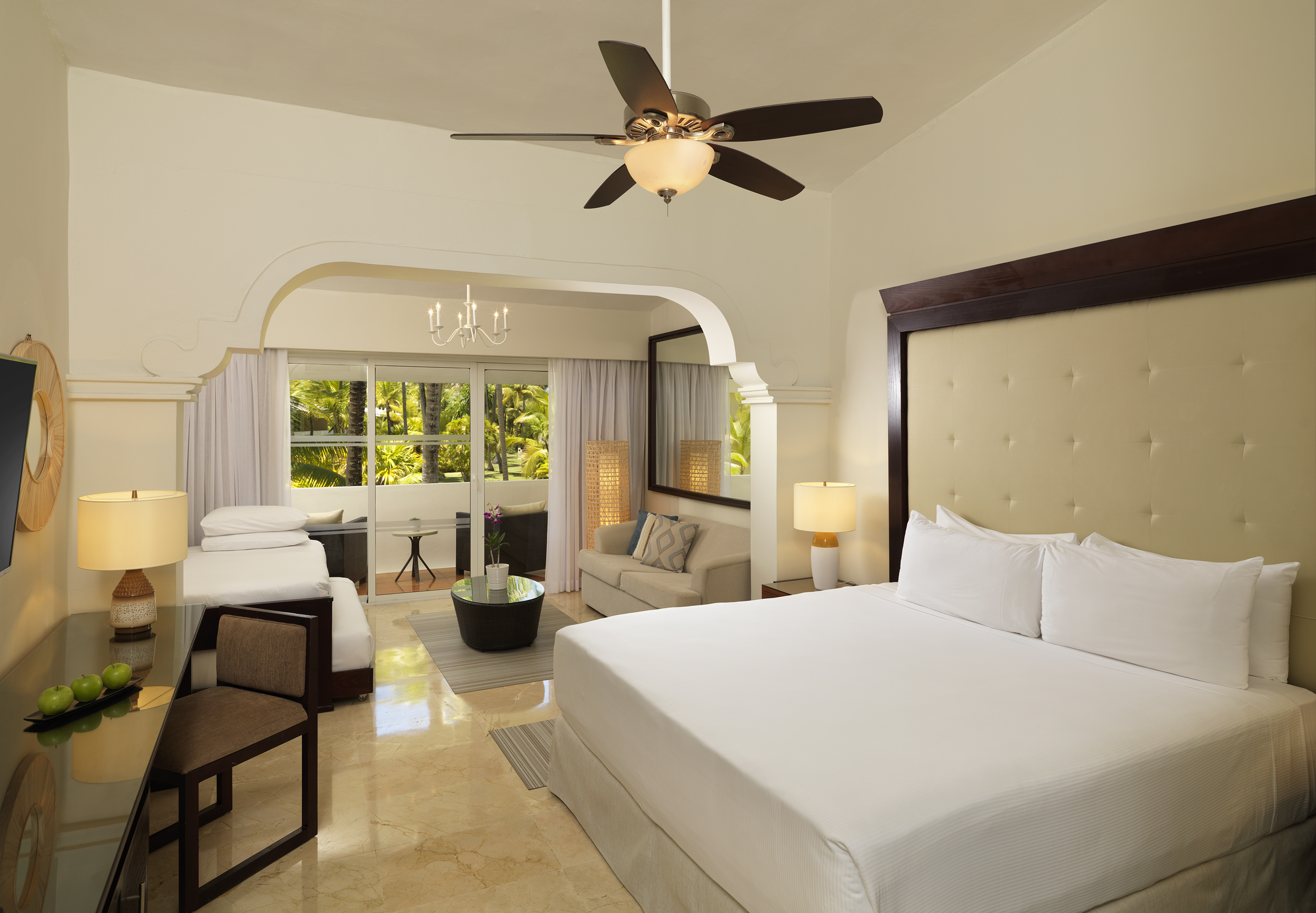 Resort hotel room with king bed, sofa, tropical balcony, and ceiling fan.