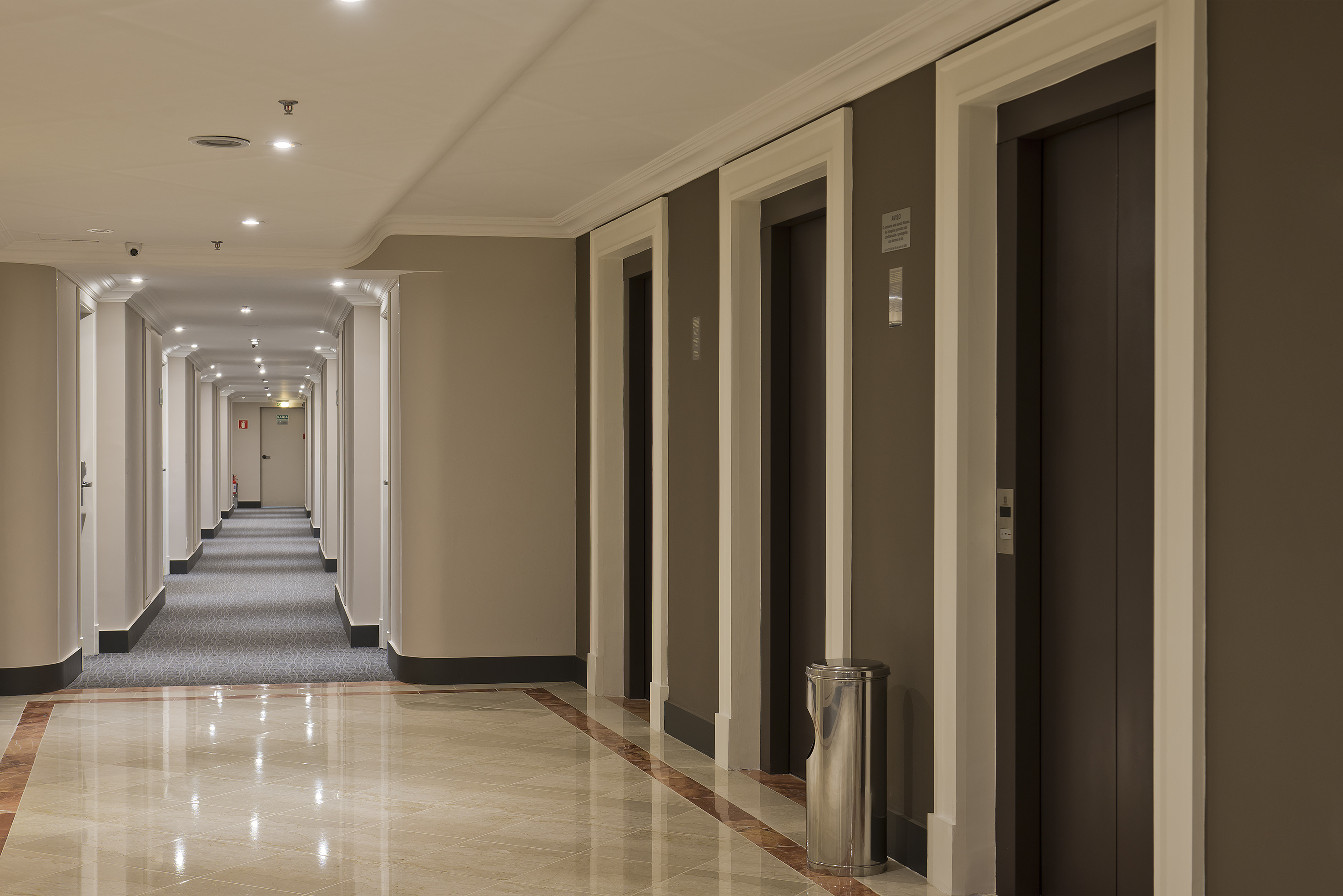 Hotel corridor with dark elevator doors, shiny tiled floor, and long hallway.