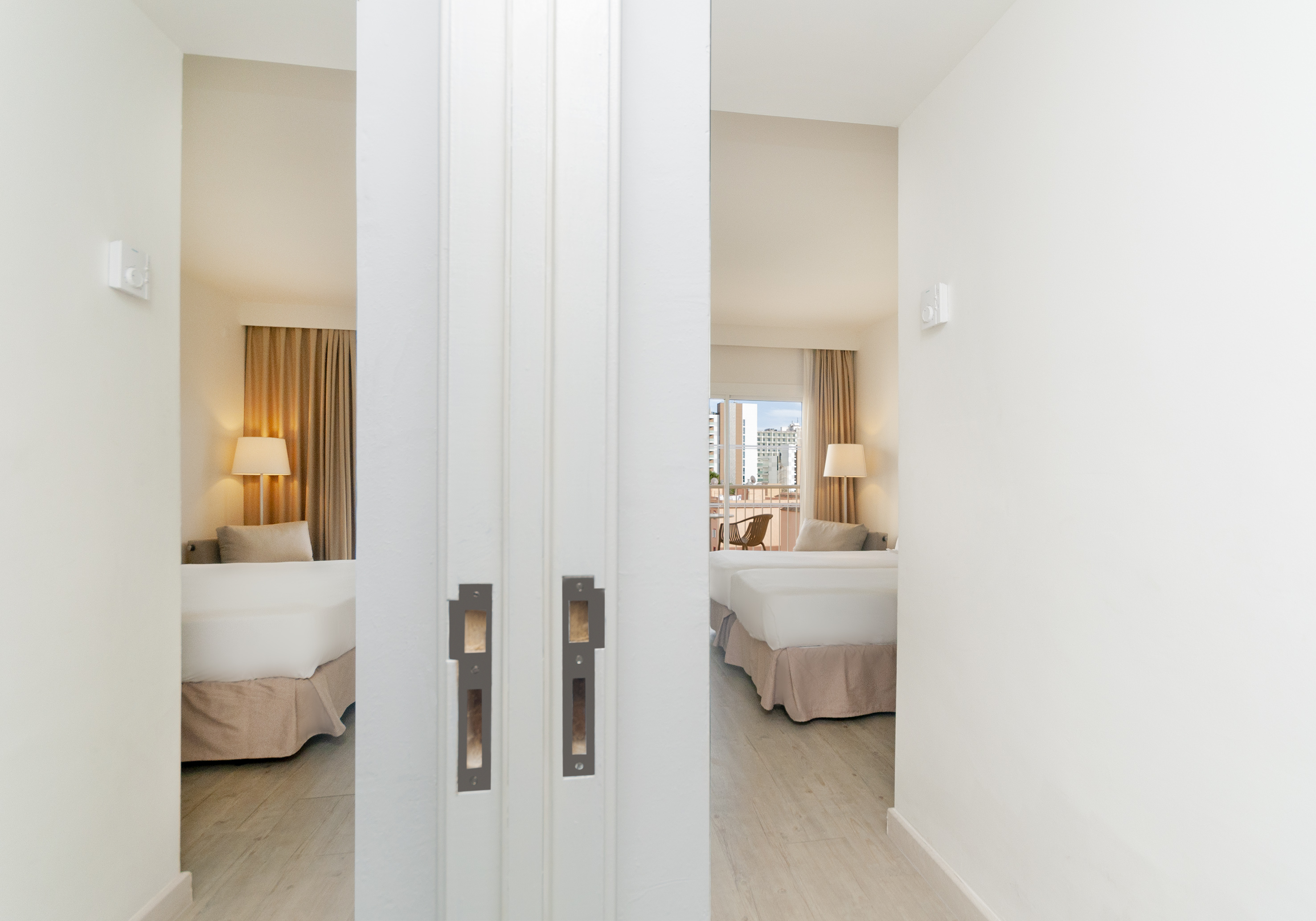 Hallway view into a bright hotel room with twin beds and balcony access.