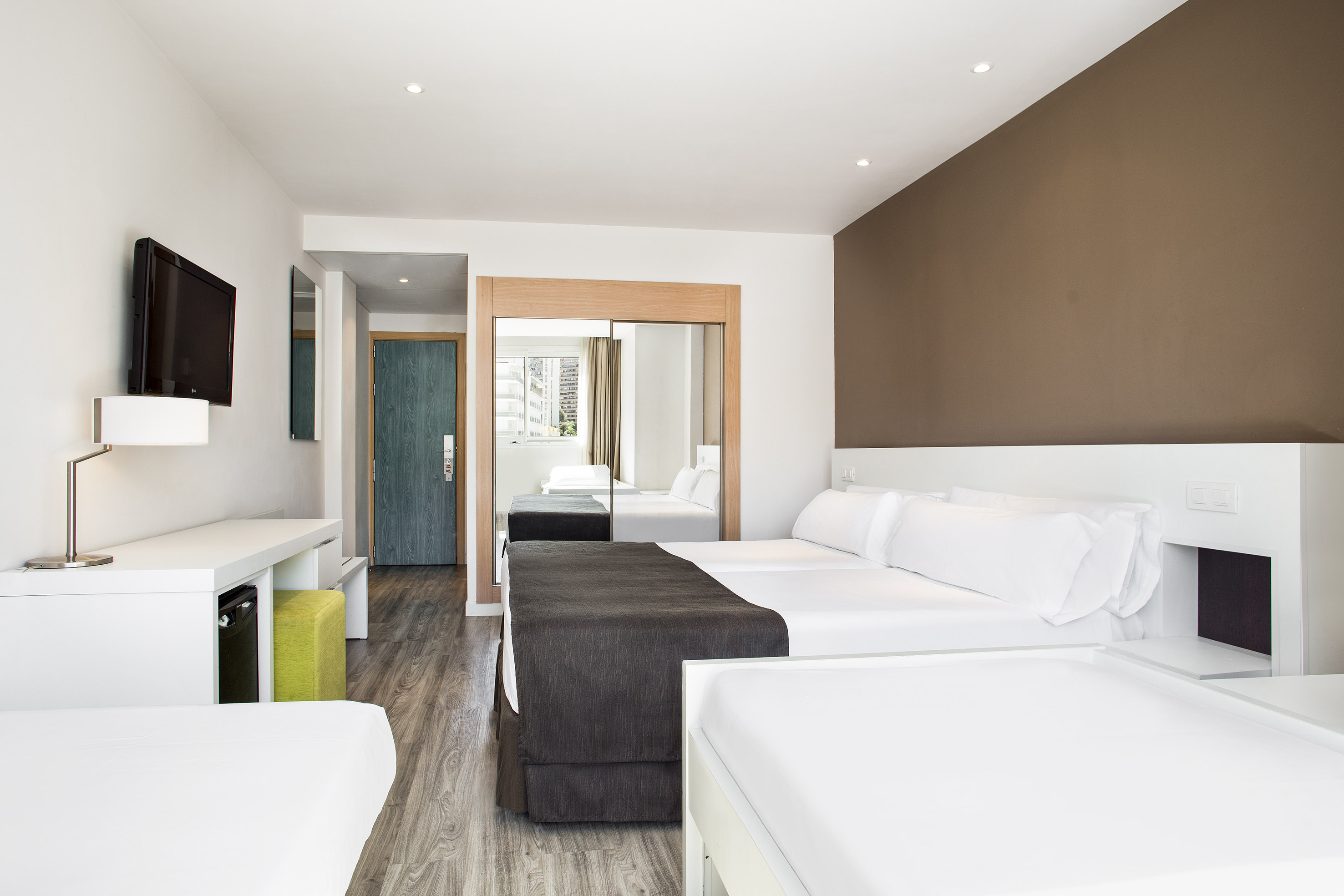 Modern triple guest room with white beds, TV, desk, and mirrored closet.
