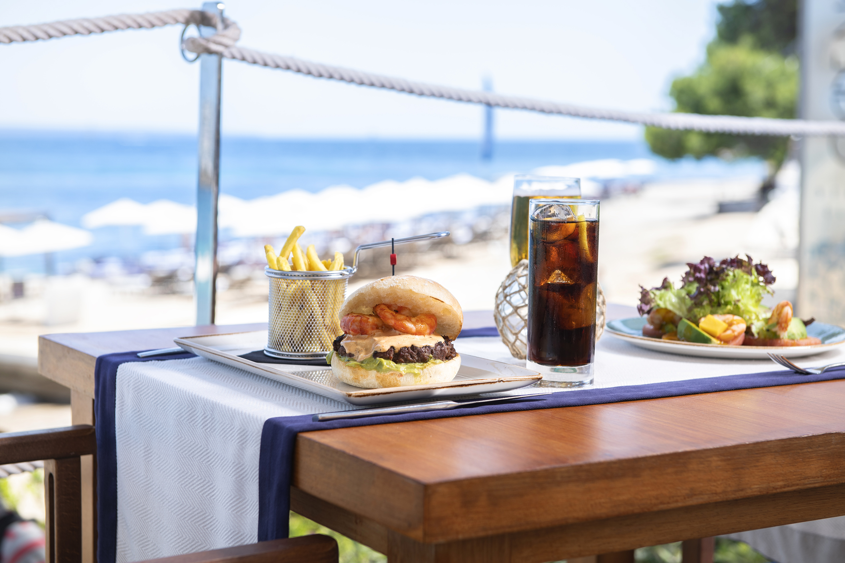 Shrimp burger, fries, and cola on an outdoor table overlooking the ocean.