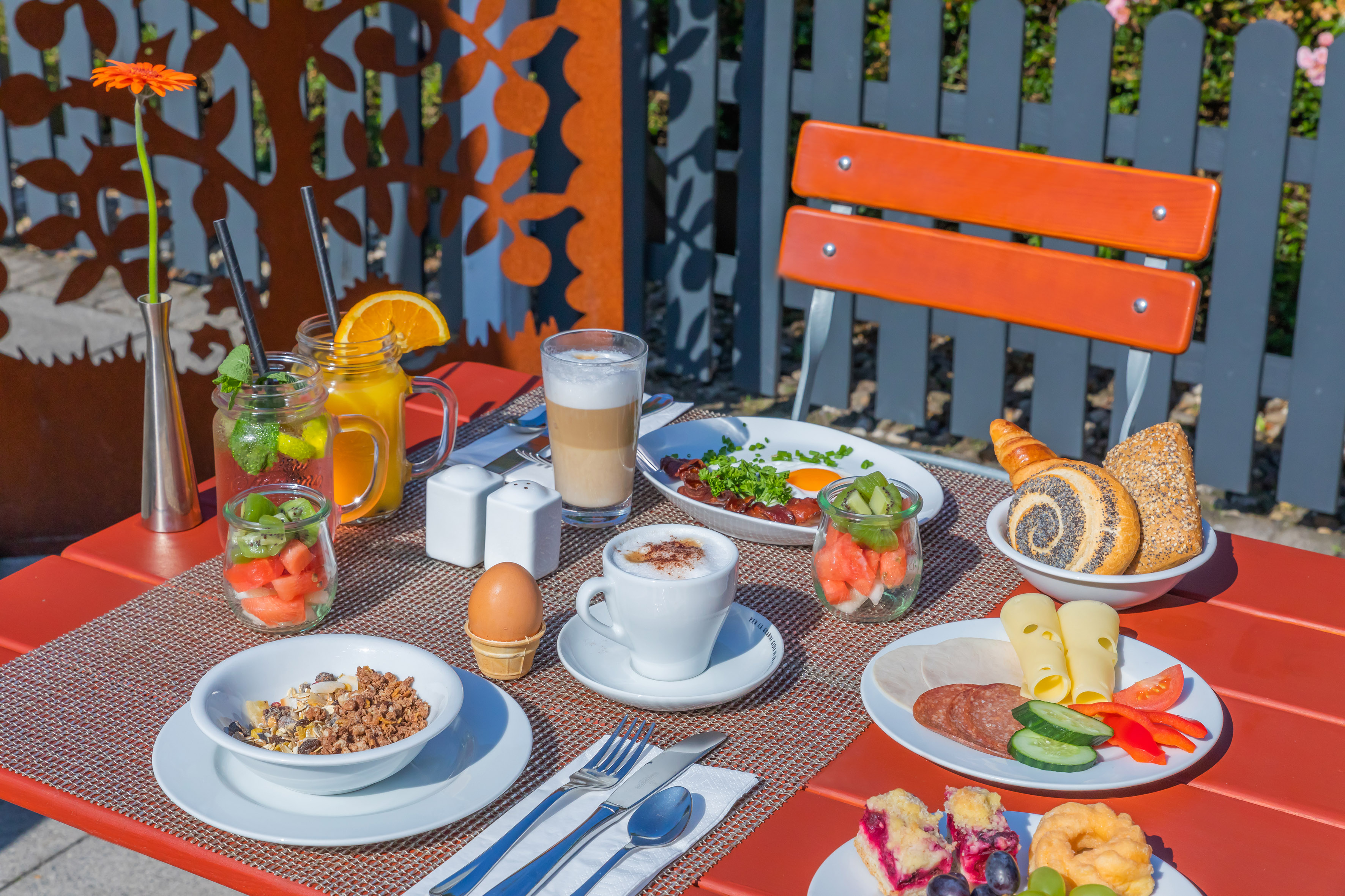 Sunny outdoor breakfast on a red table: eggs, fruit, granola, pastries, coffee.