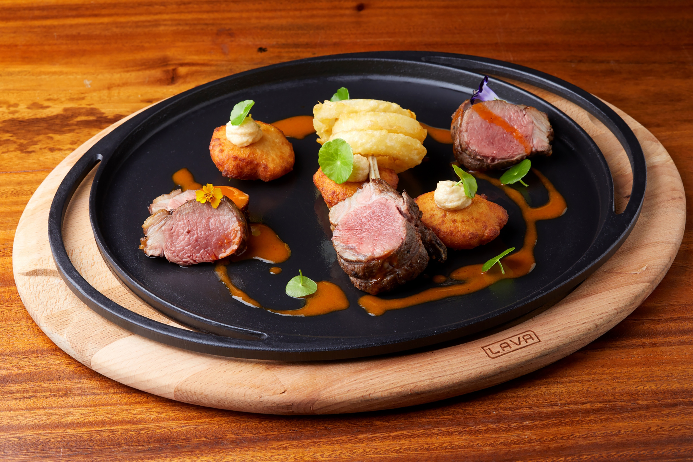 Gourmet seared lamb, croquettes, onion rings, and sauce on cast iron platter.
