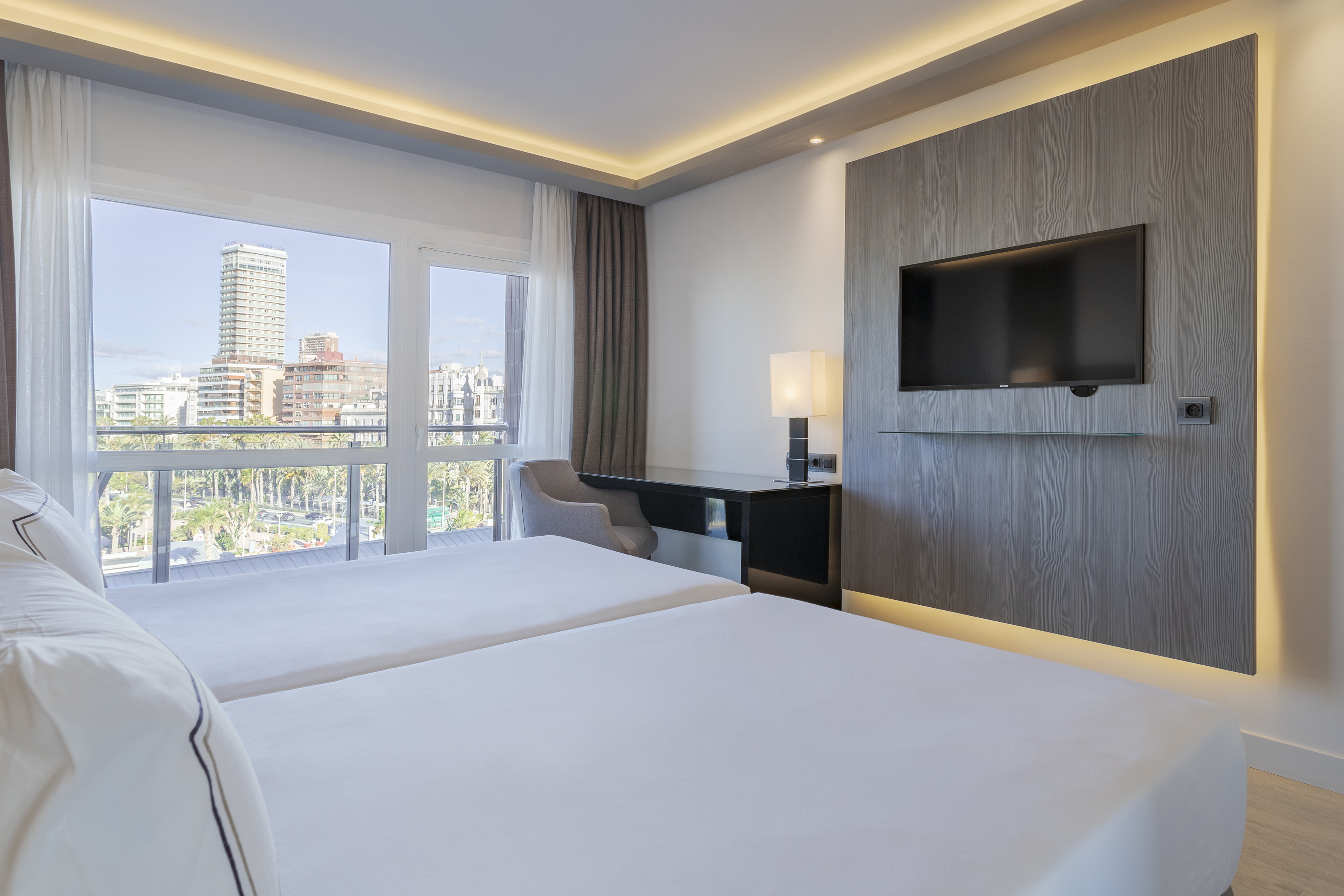Modern hotel room with twin beds, city view, desk area, and wall-mounted TV.