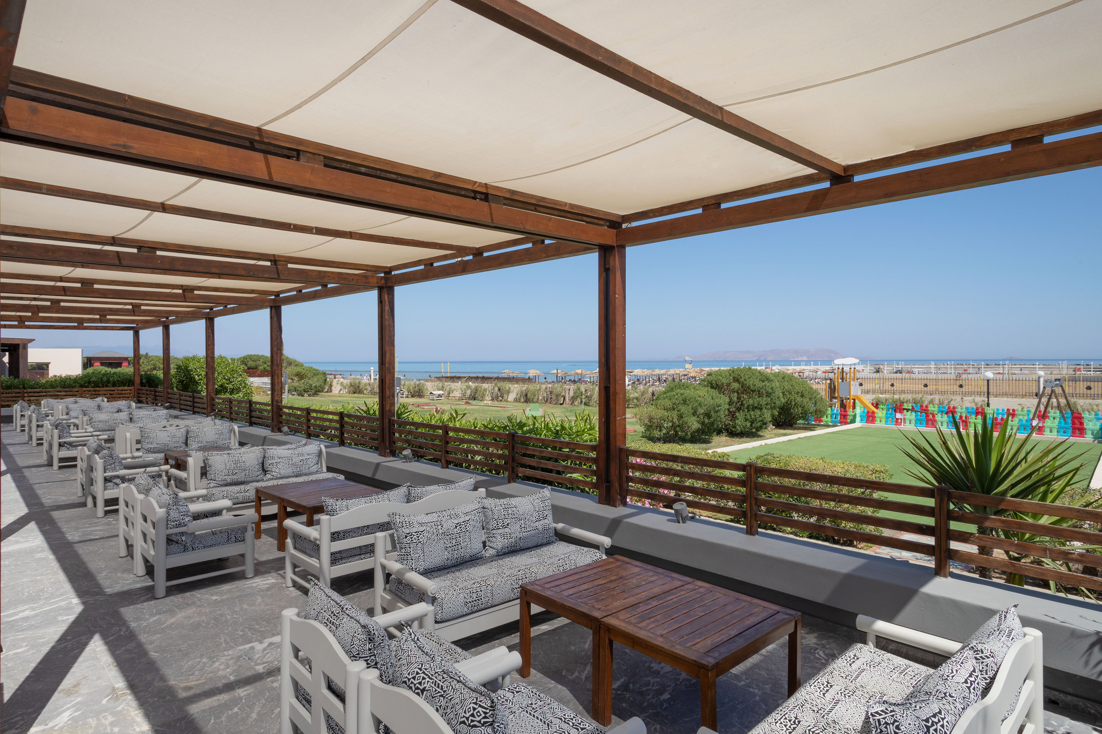 Hotel patio with shaded seating, ocean view, green lawn, and kids' playground.