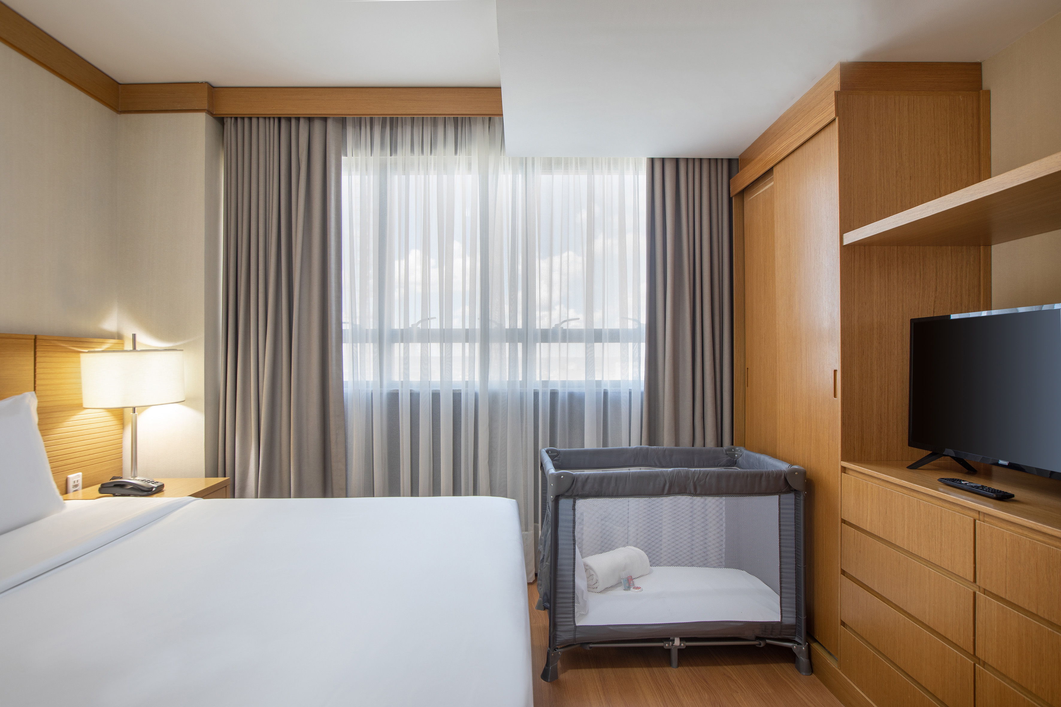 Modern family-friendly hotel room with bed, baby crib, and bright window.