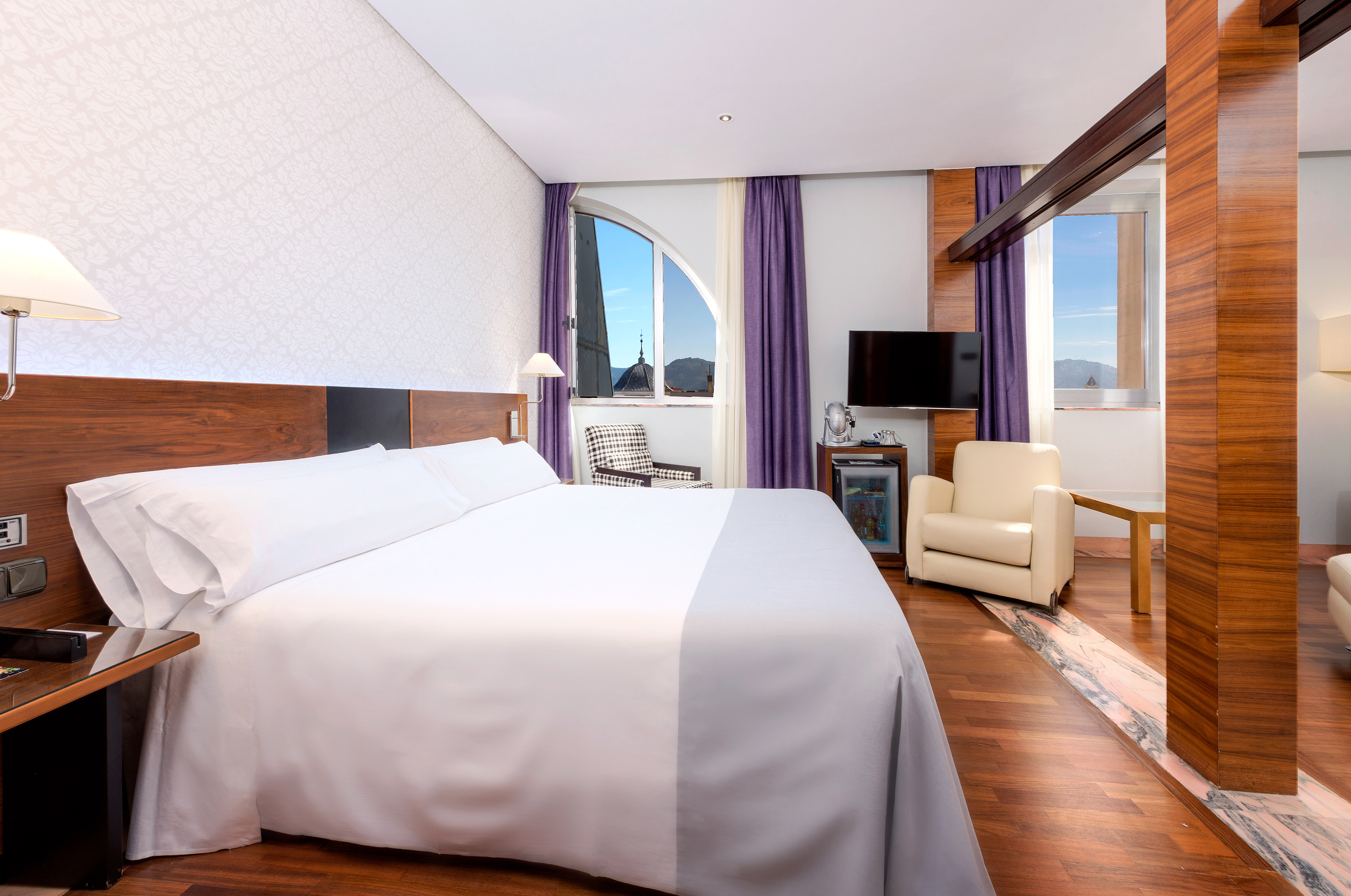 Modern hotel room with king bed, arched window city view, wood accents.