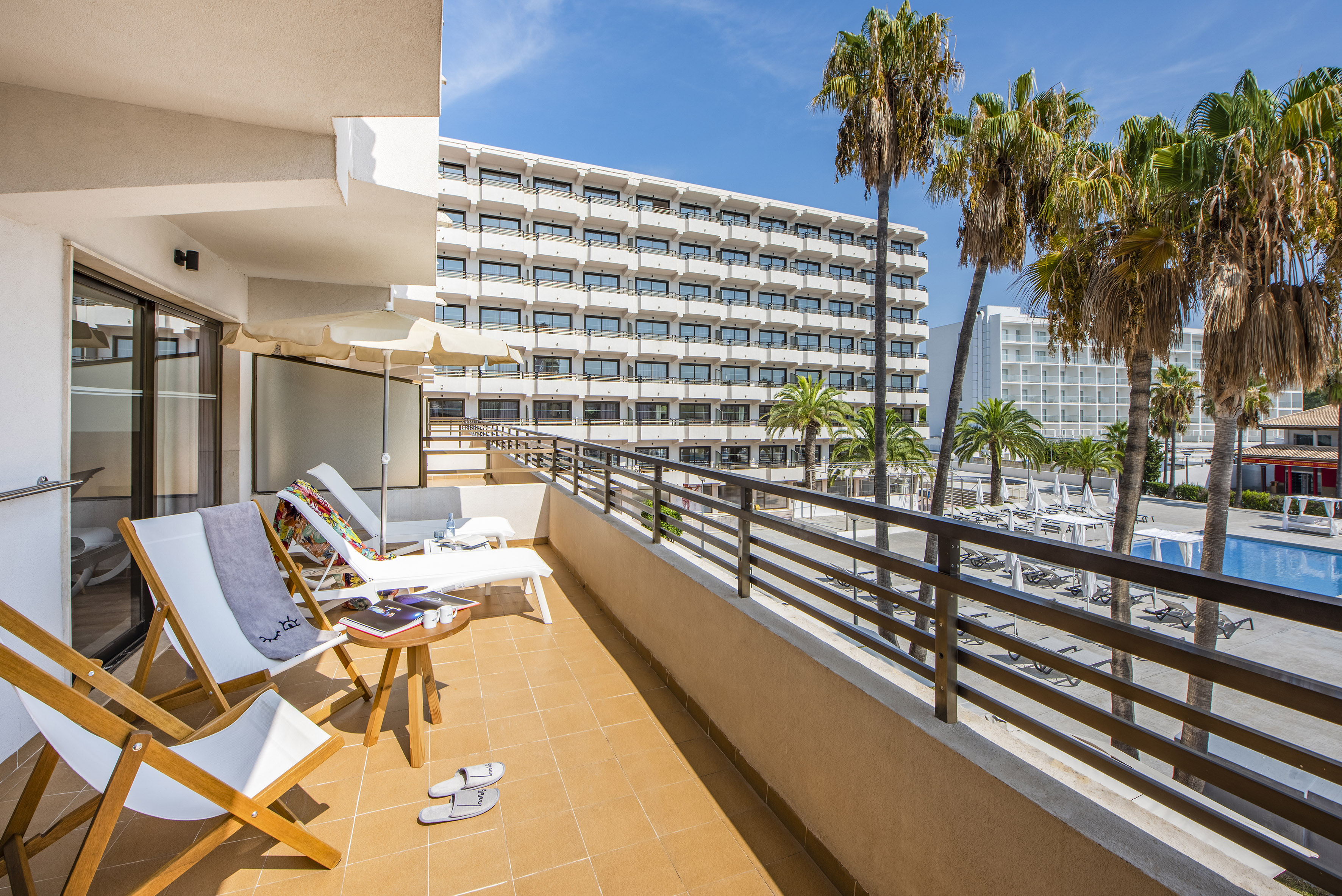 Sunny hotel balcony overlooking resort pool and palm trees.