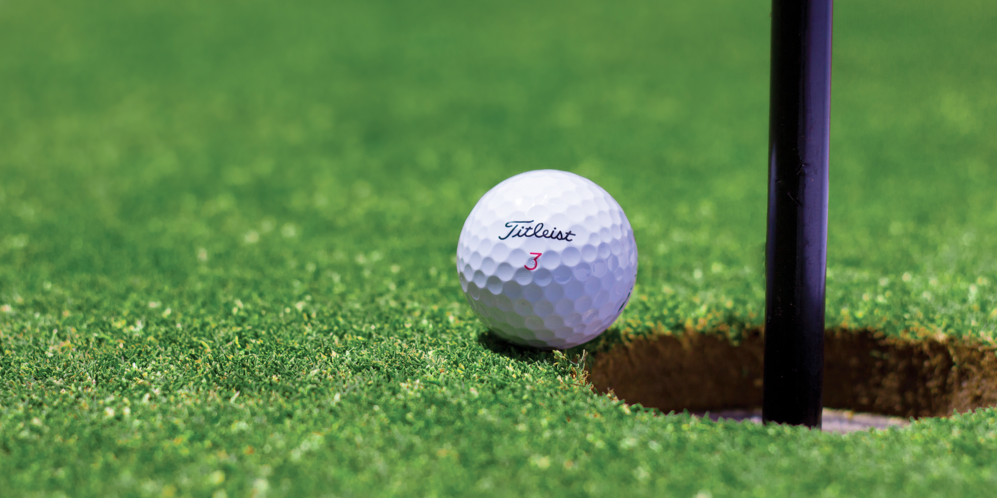 White Titleist 3 golf ball next to golf hole and flagstick on putting green.