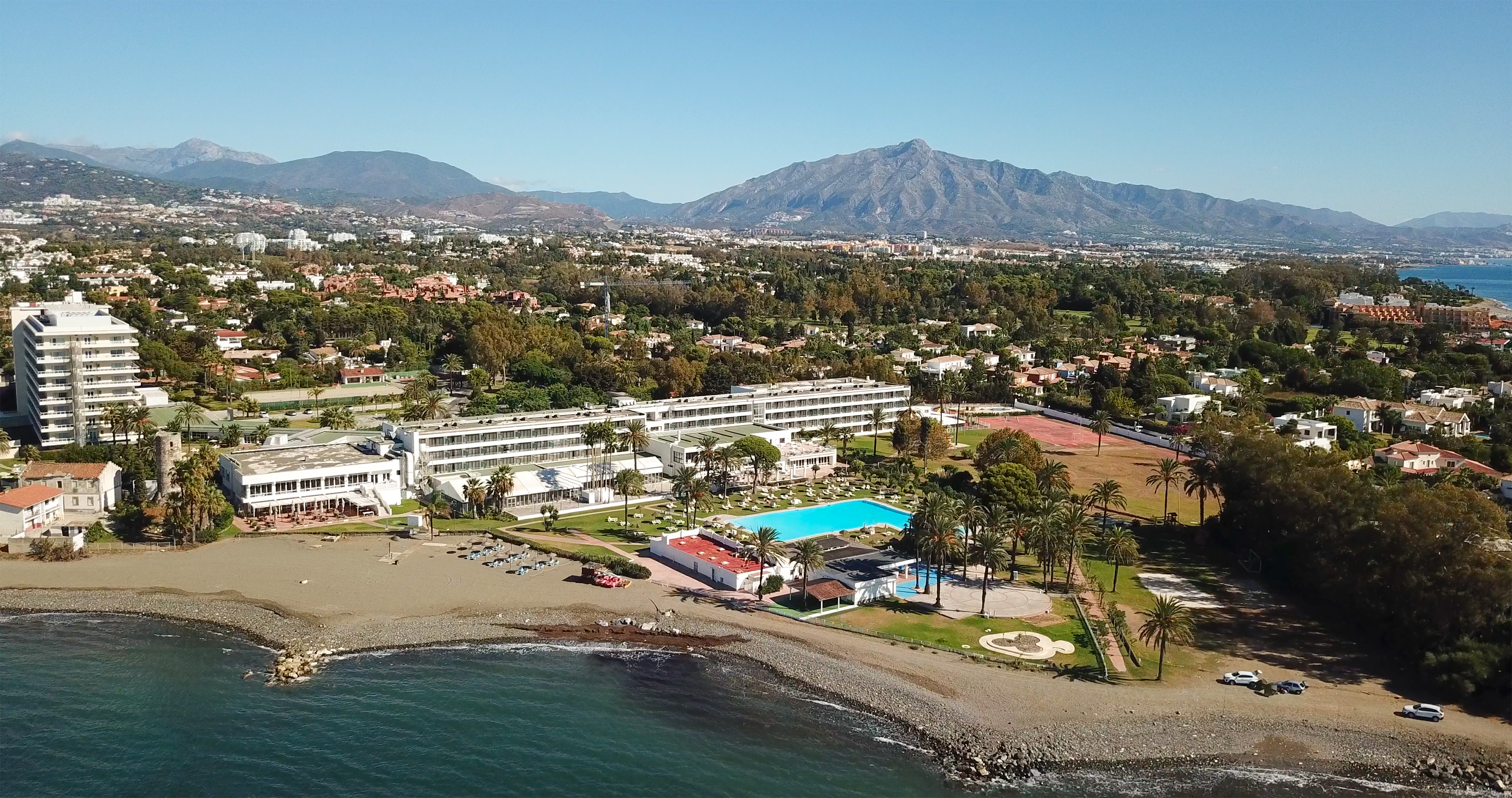 Aerial view of beachfront resort with large pool, palm trees, sea, and mountains.