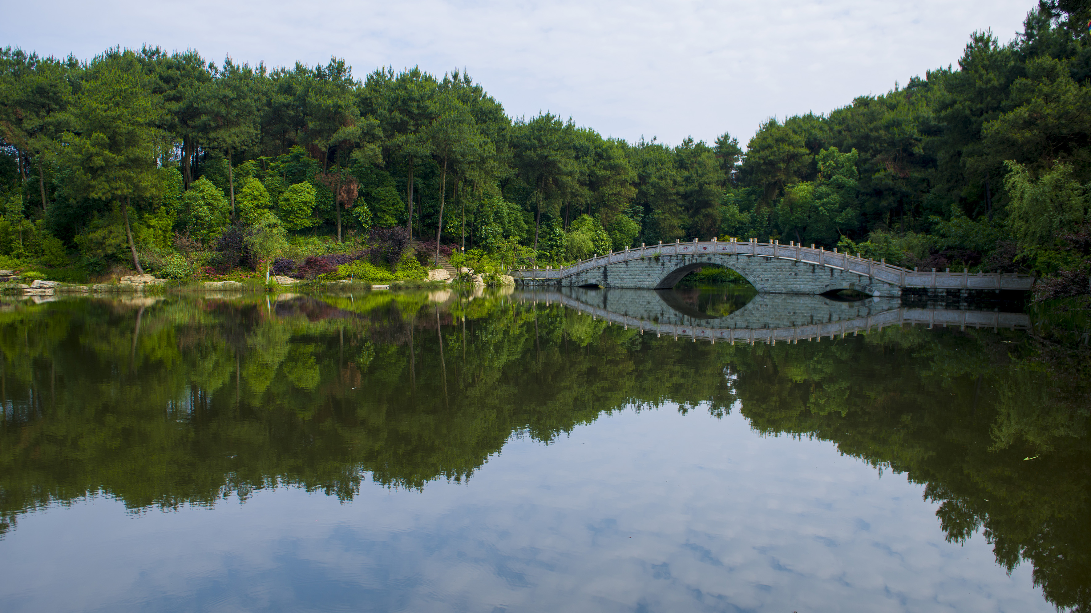 Traditional stone arch bridge over calm water, lush forest reflections.