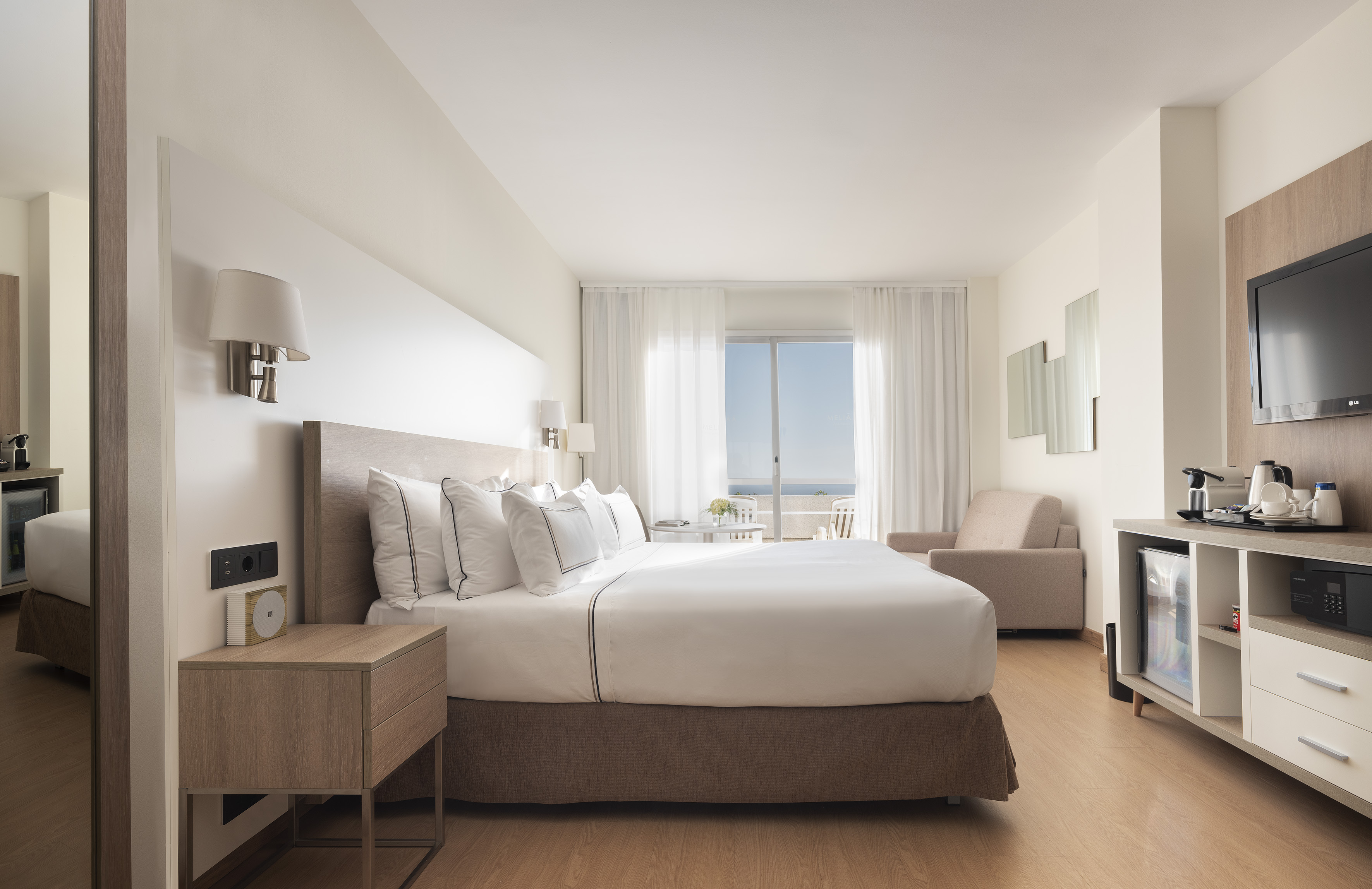Modern hotel bedroom with king bed, ocean view balcony, and flat-screen TV.