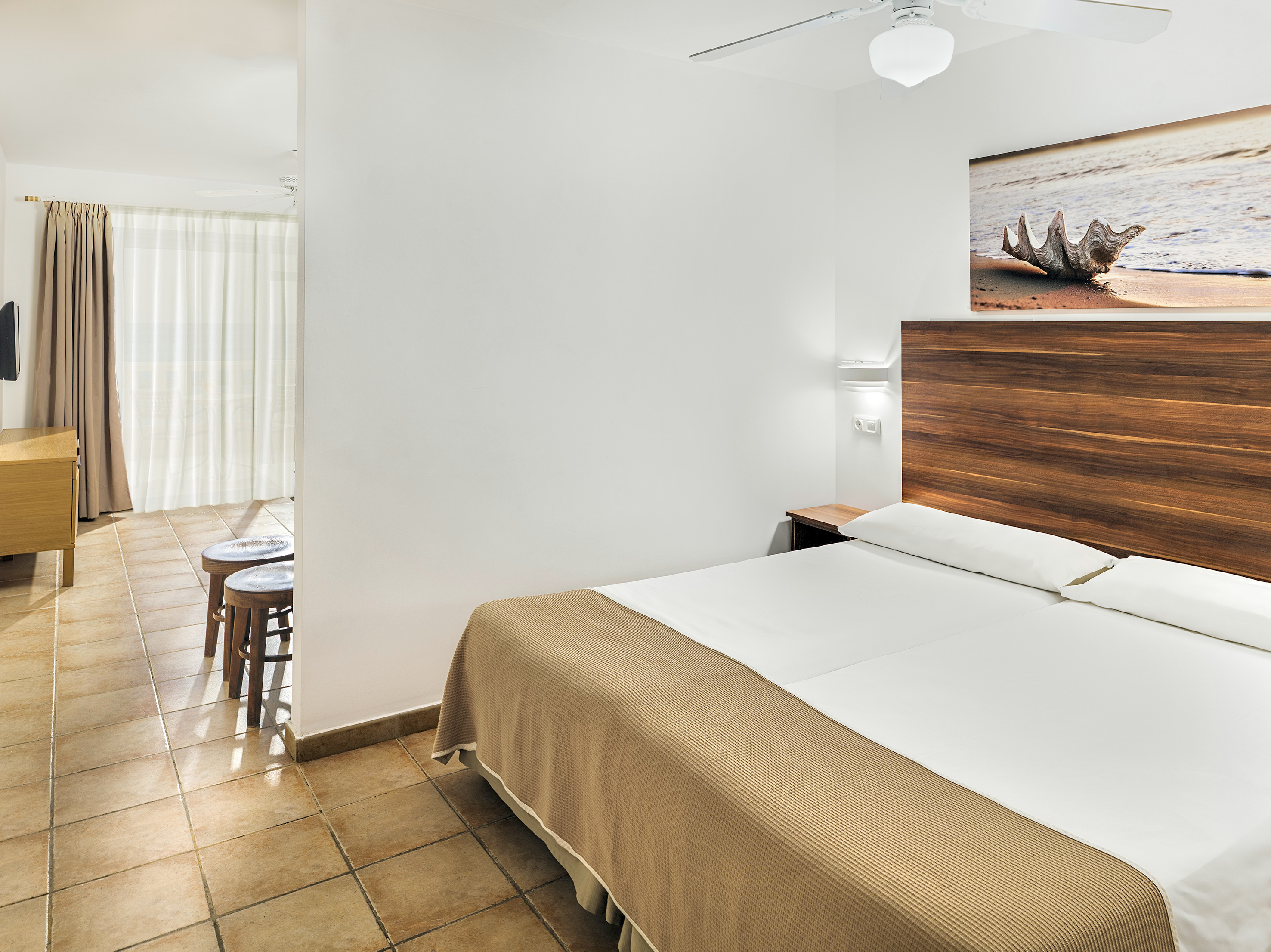 Modern hotel bedroom with double bed, wooden headboard, beach art, tiled floor.