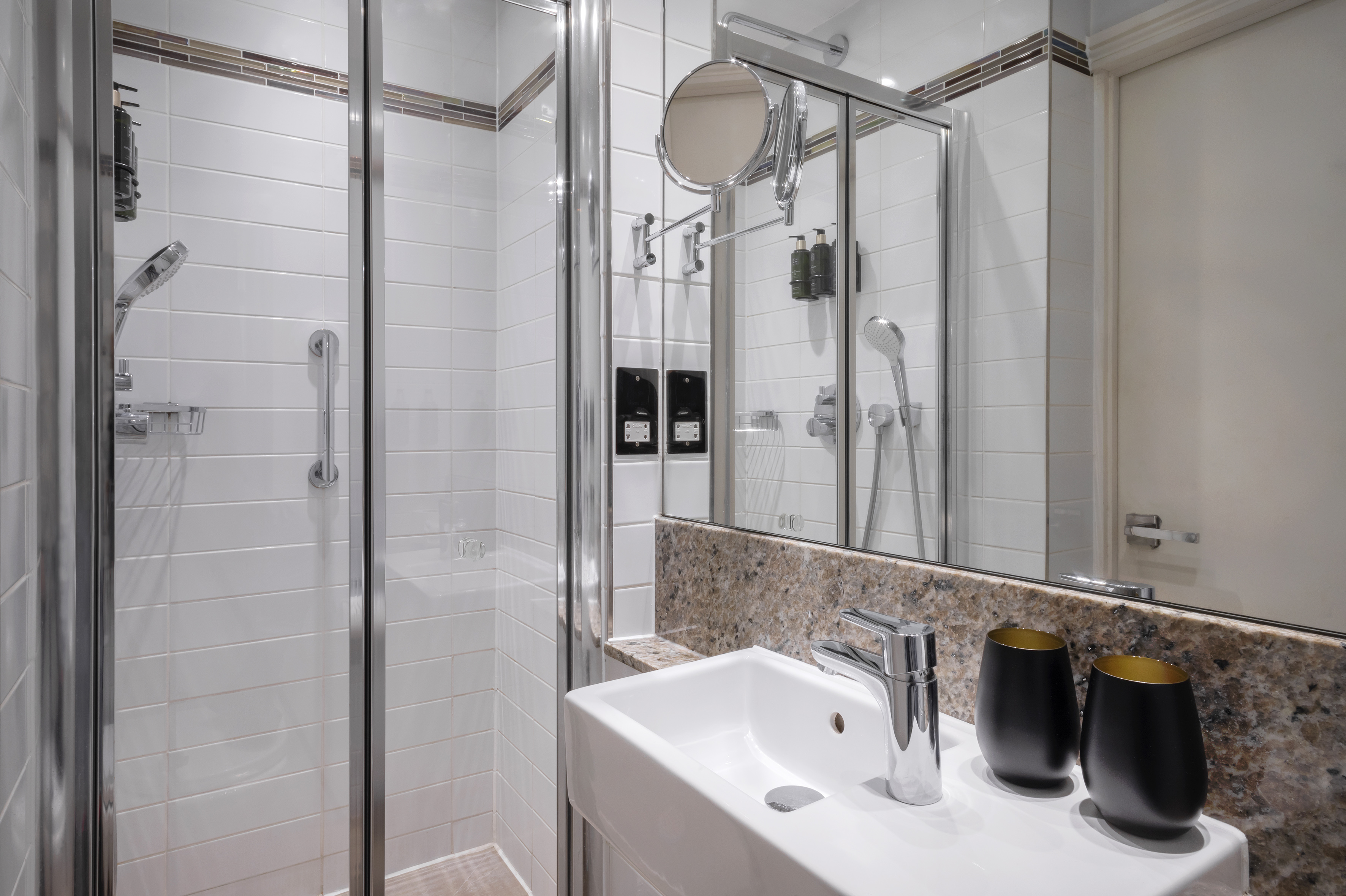 Modern hotel bathroom with walk-in shower, white tiles, sink, and granite counter.