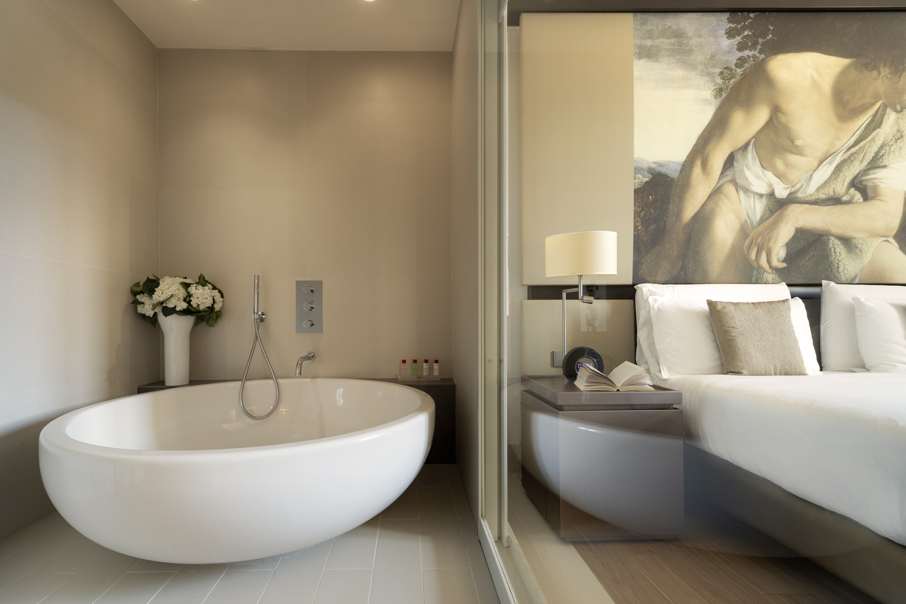 Modern hotel bathroom with freestanding tub and glass-walled bedroom.