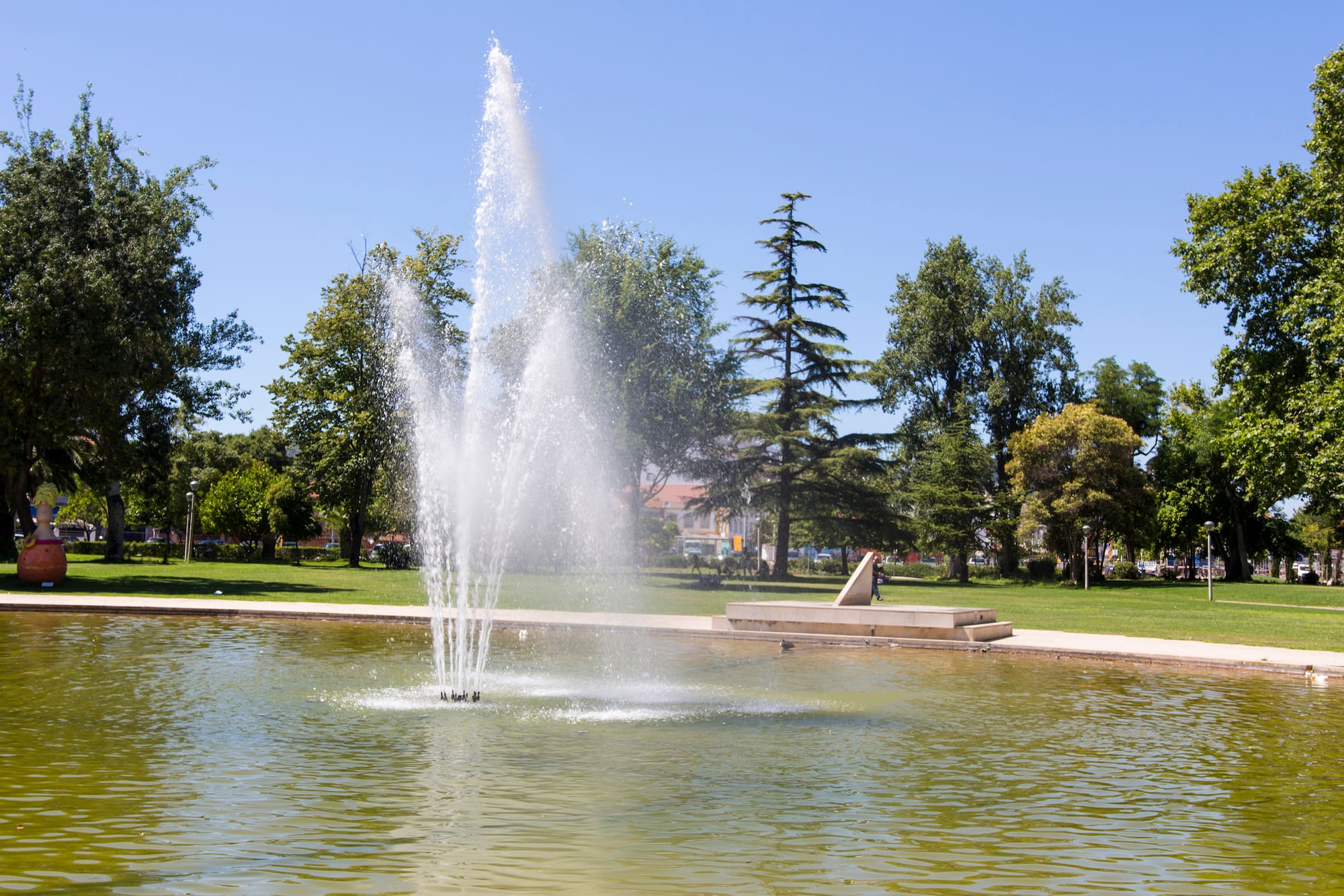 Fountain spraying water in a park pond surrounded by green trees under a blue sky.