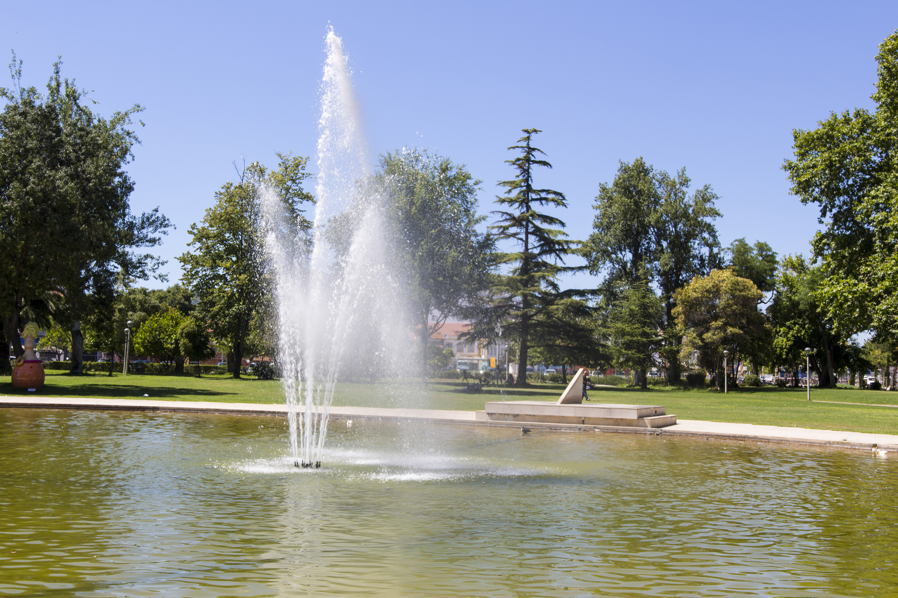 Fountain spraying water in a park pond surrounded by green trees under a blue sky.