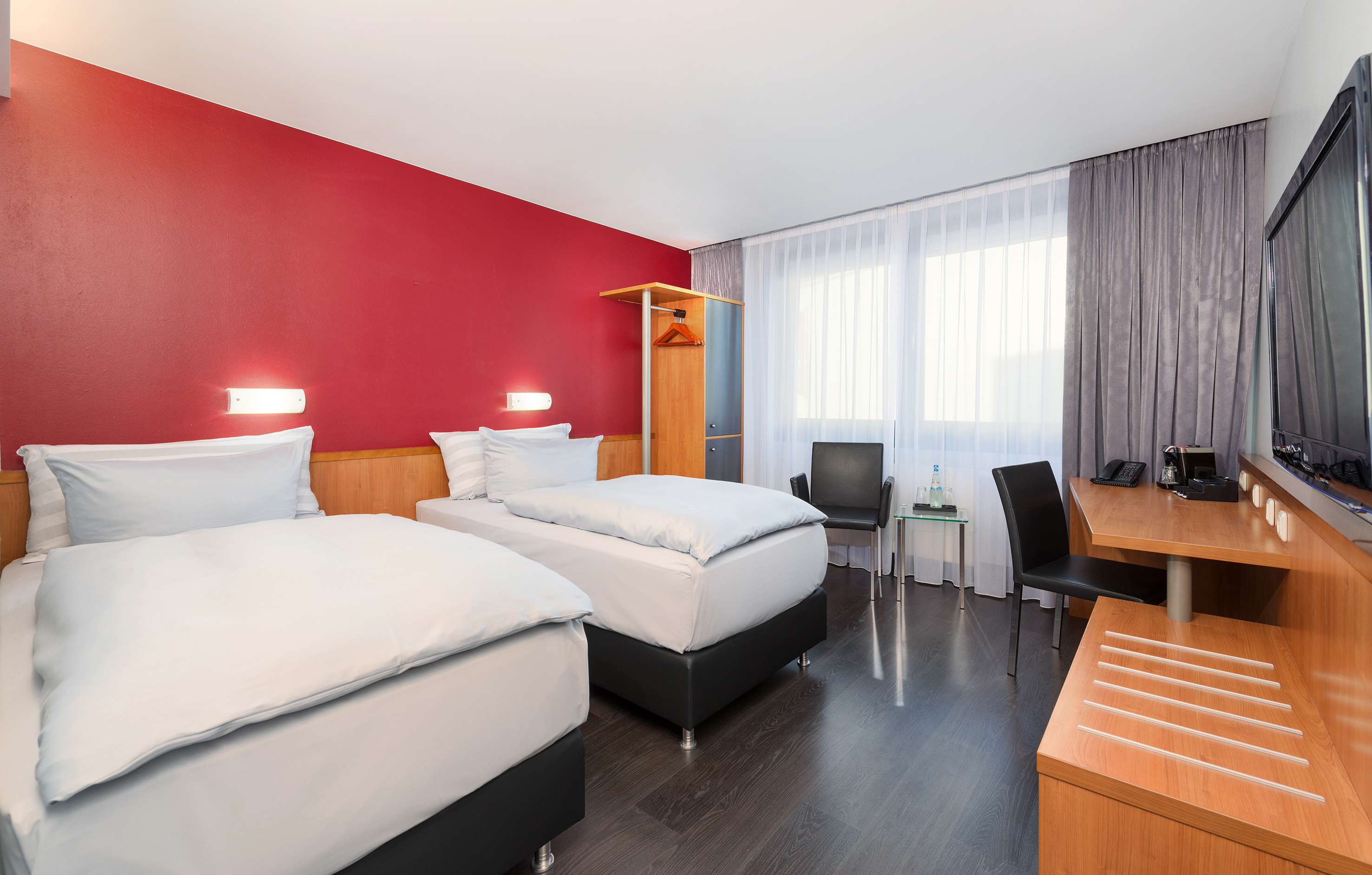 Modern hotel room with twin beds, red accent wall, desk, and large window.