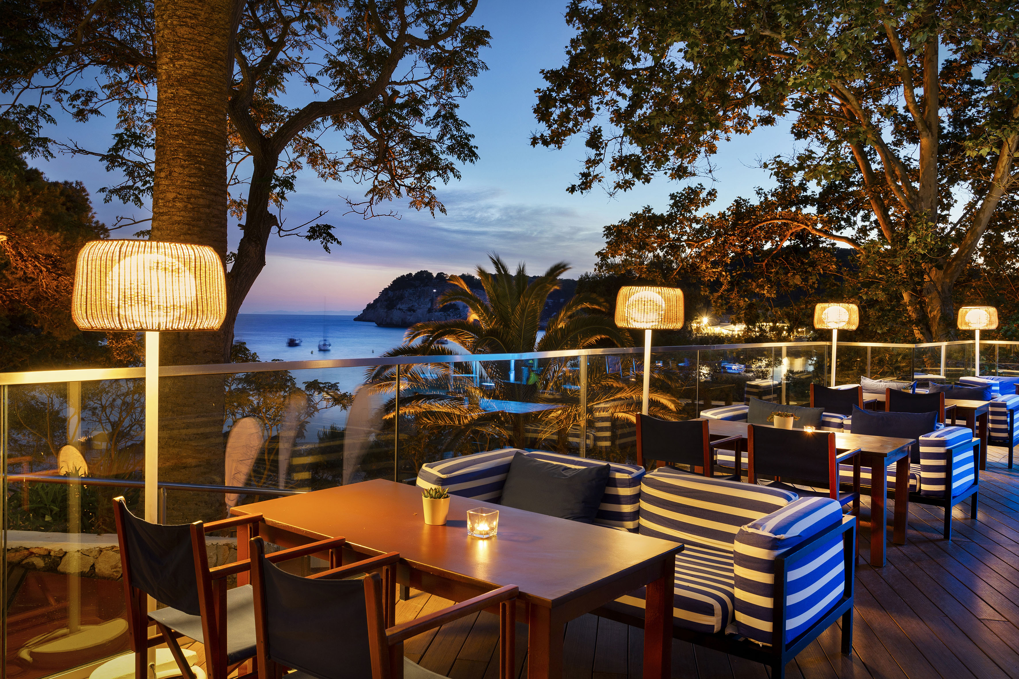 Dusk restaurant terrace with bay view, warm lights, and blue striped seating.